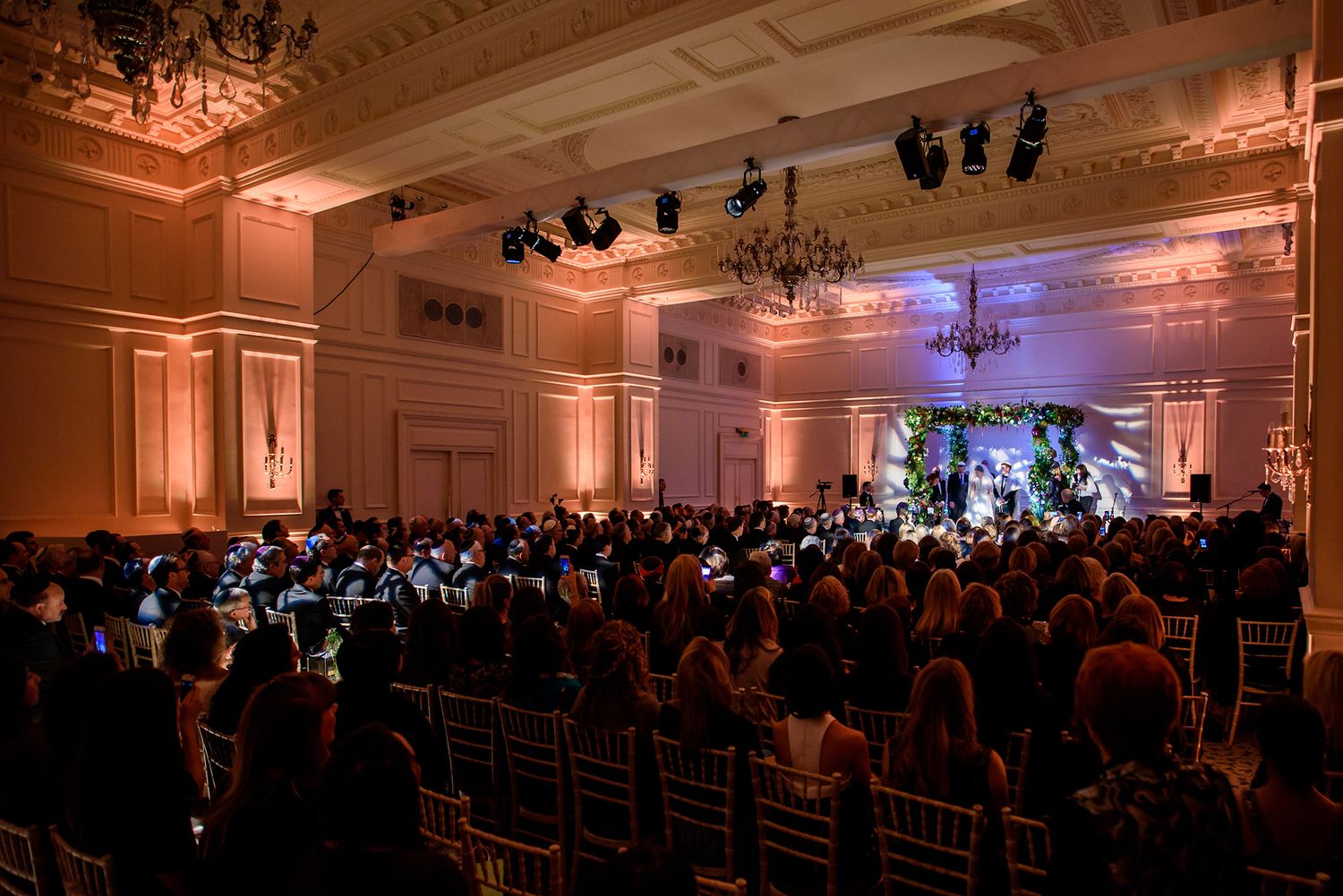 Large elegant ballroom with ornate chandeliers and crown molding filled with guests for a special event.