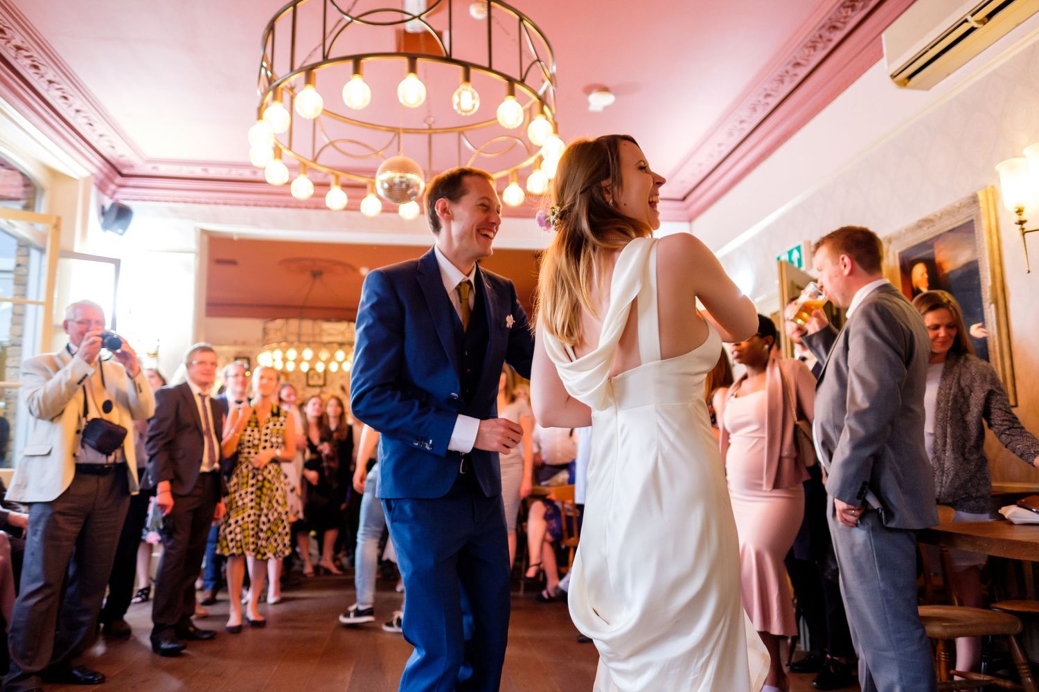 Couple dances at wedding reception while guests watch in pink-lit room with chandelier.