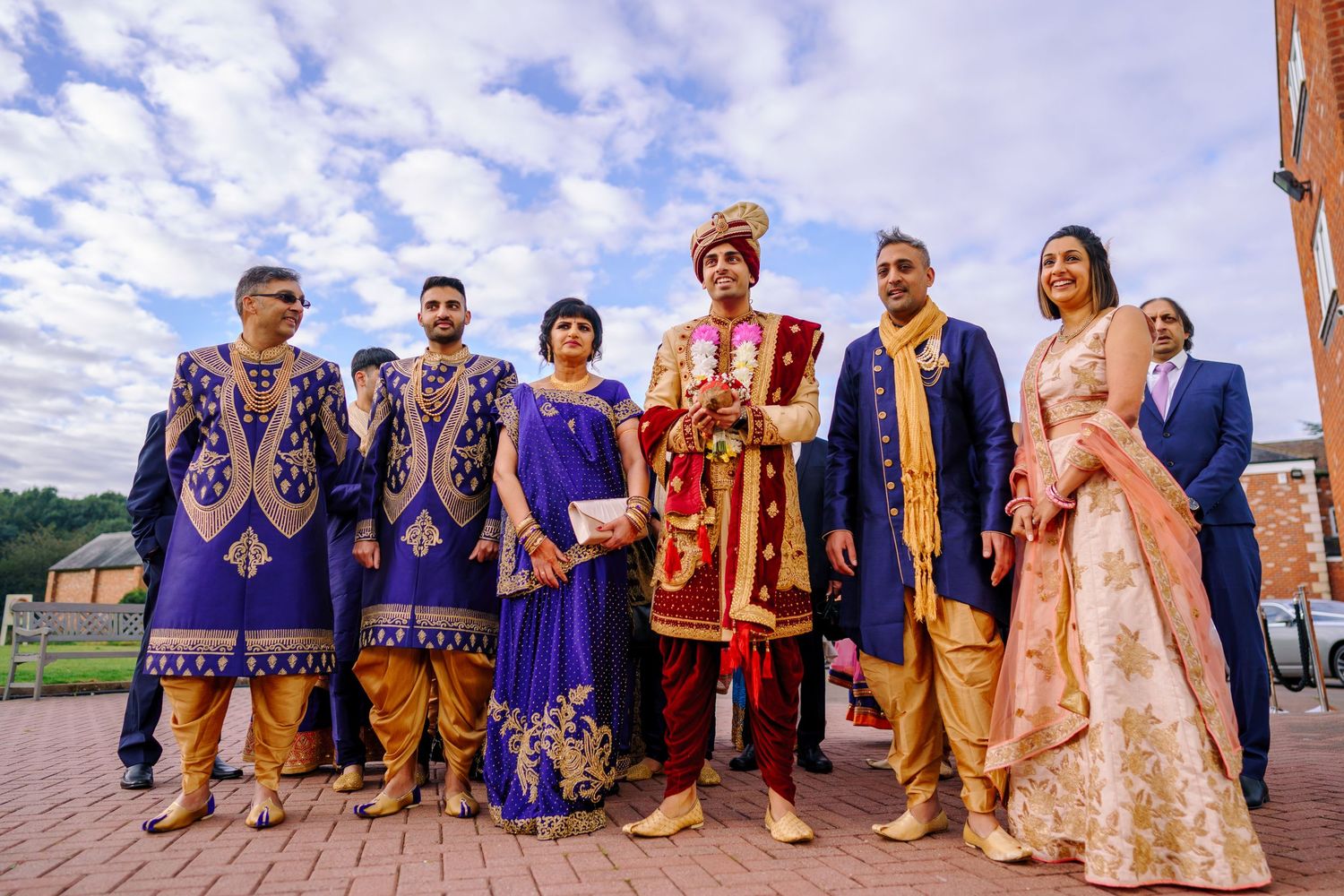 Wedding party poses outdoors in traditional Indian formal wear with blue sky backdrop.