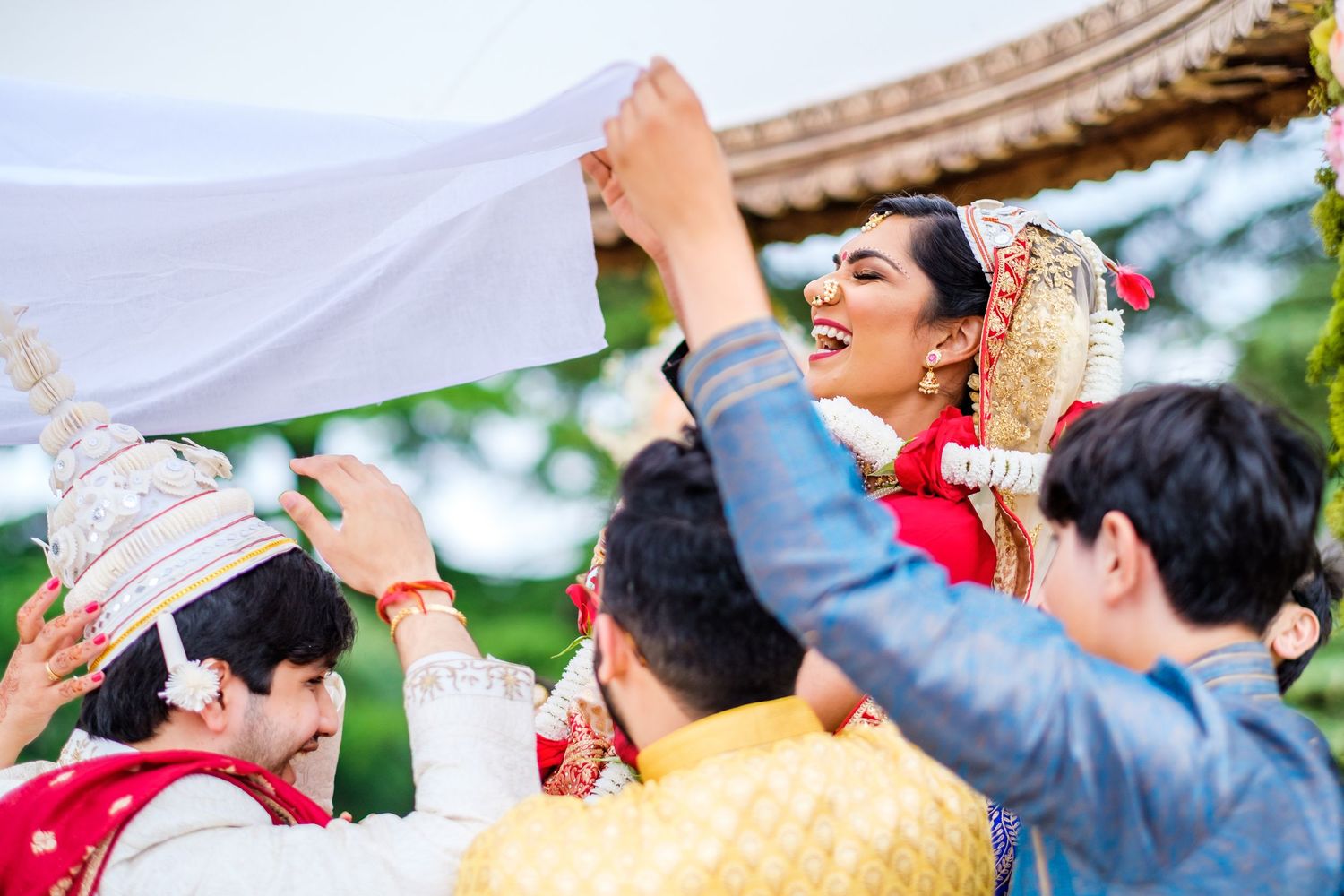 Celebratory moment during Indian wedding ceremony as participants hold white fabric overhead.