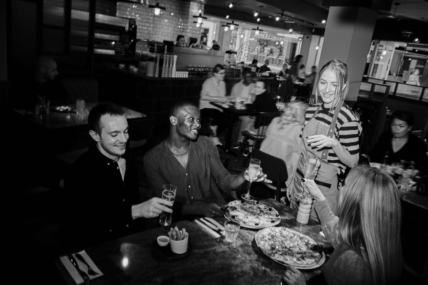 Restaurant patrons enjoying pizza and drinks in dimly lit dining room.