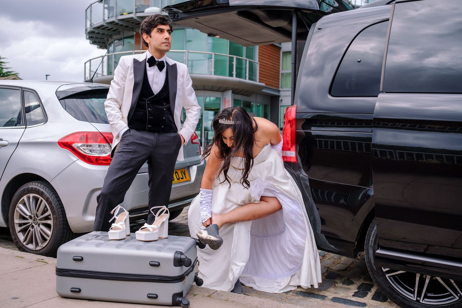 Wedding party members stand near parked cars outside a venue on an overcast day.