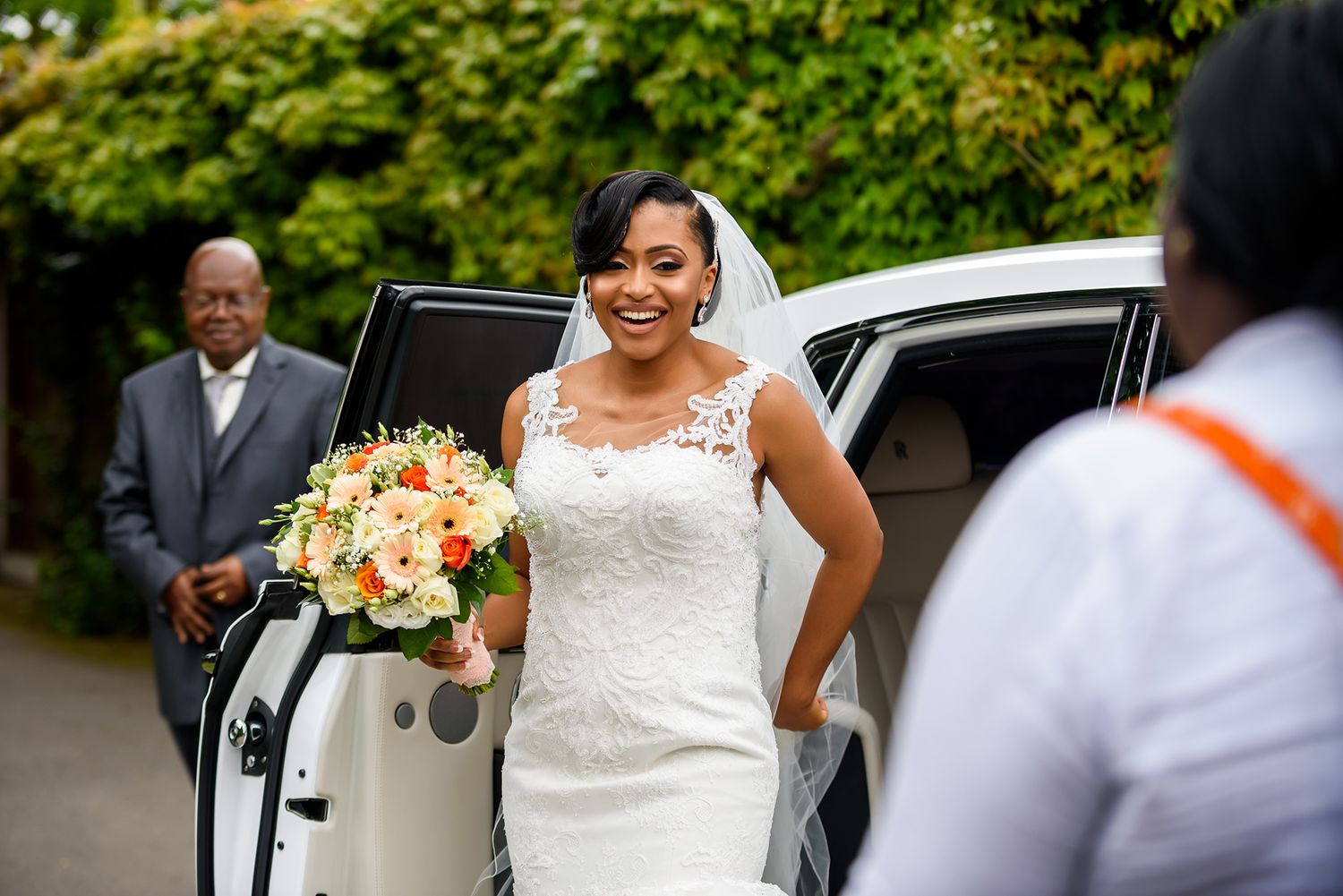 Bride in white lace dress arrives at wedding venue with floral bouquet.