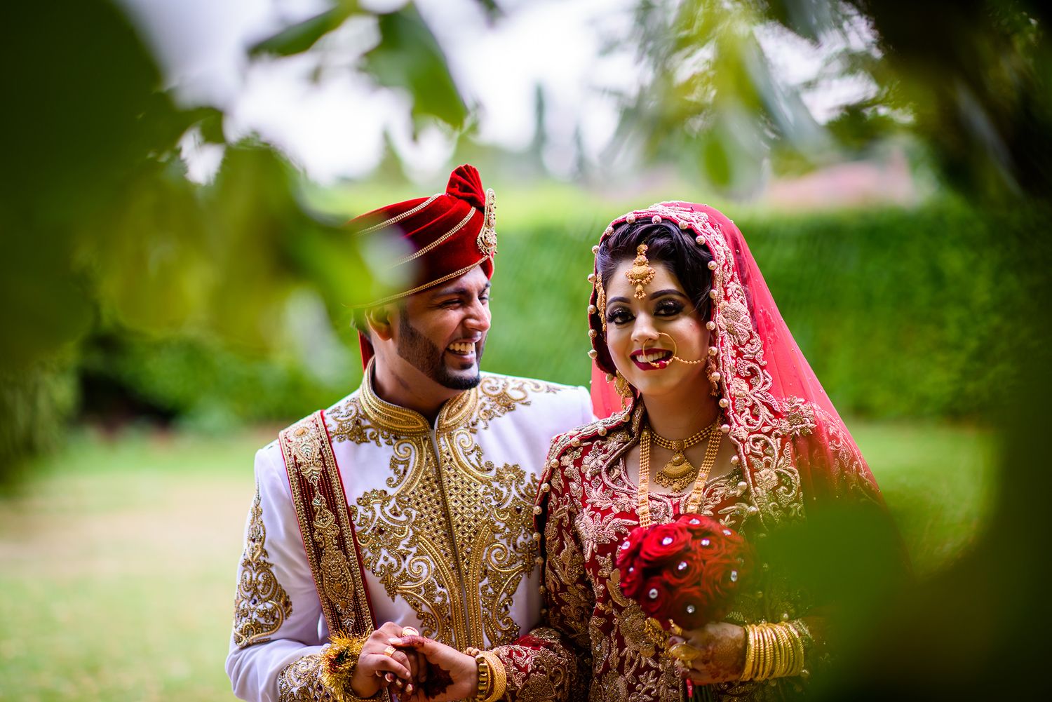 Wedding couple poses under lush green trees in traditional Indian attire during outdoor photoshoot.