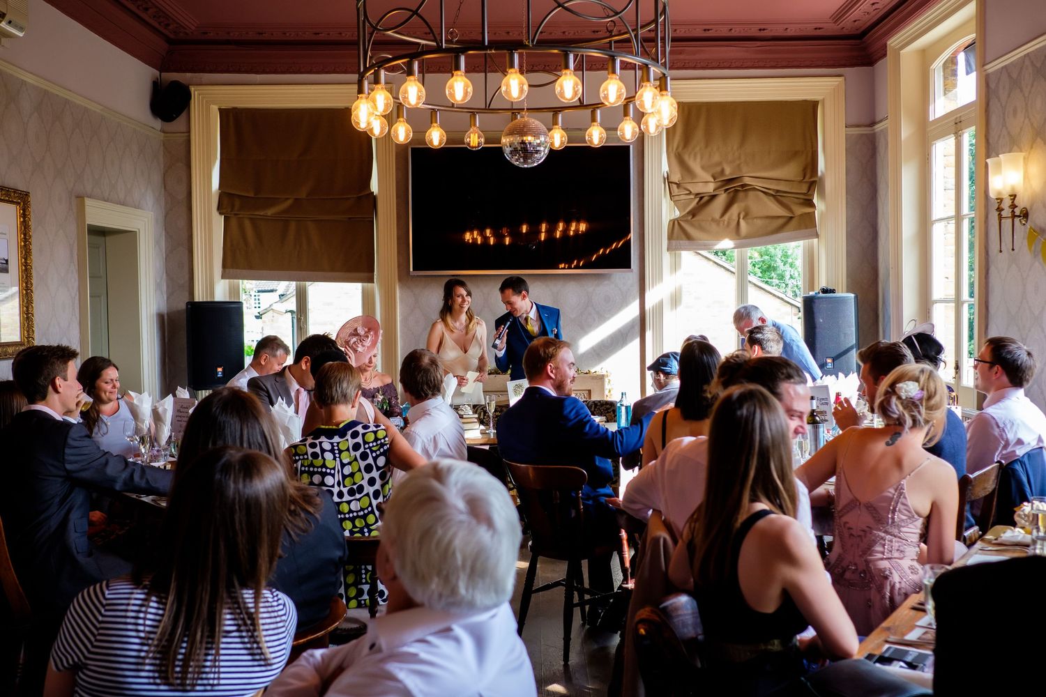 Wedding reception venue with guests seated around tables under elegant chandelier lighting.