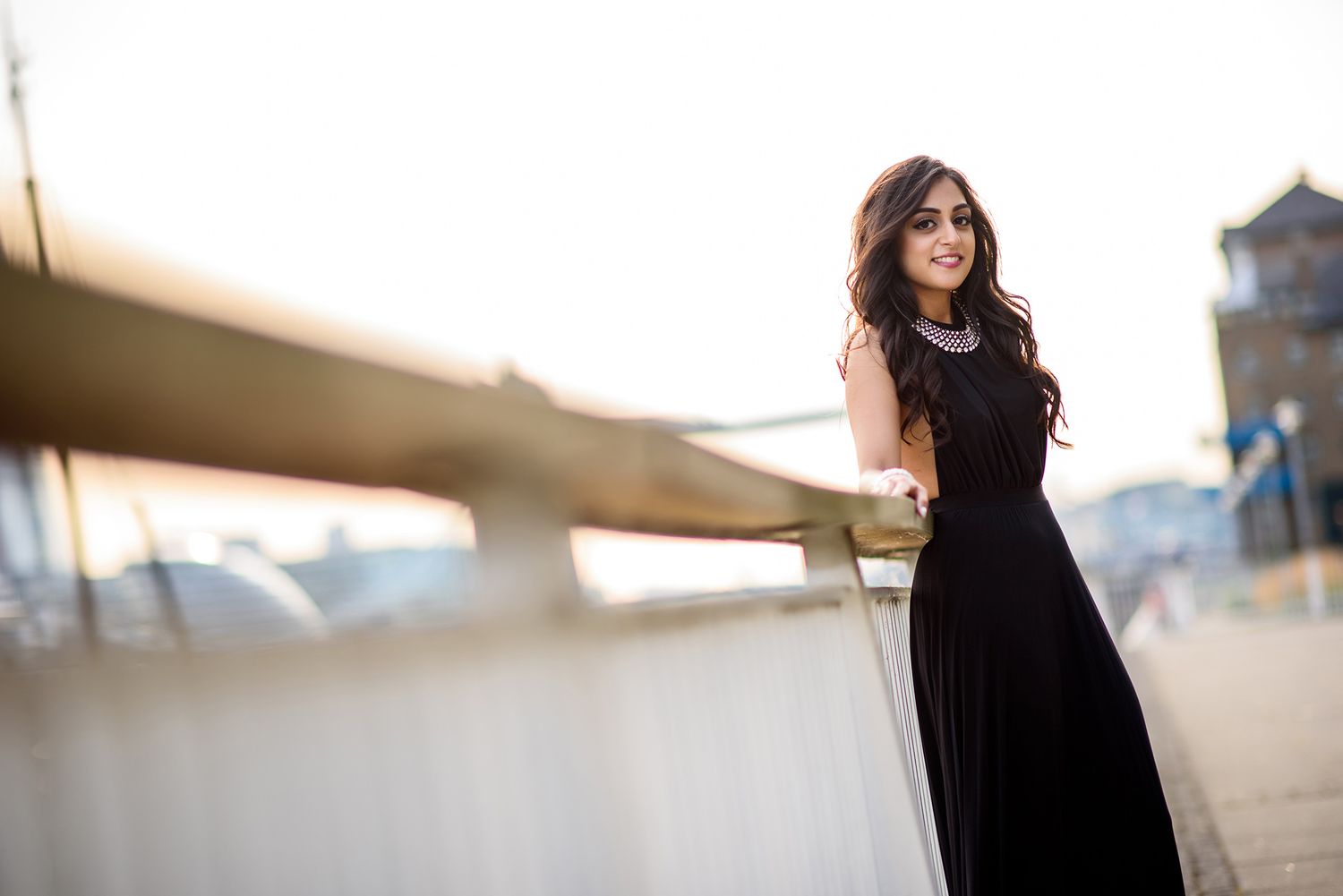 Woman in elegant black dress poses by waterfront with urban architecture in background.
