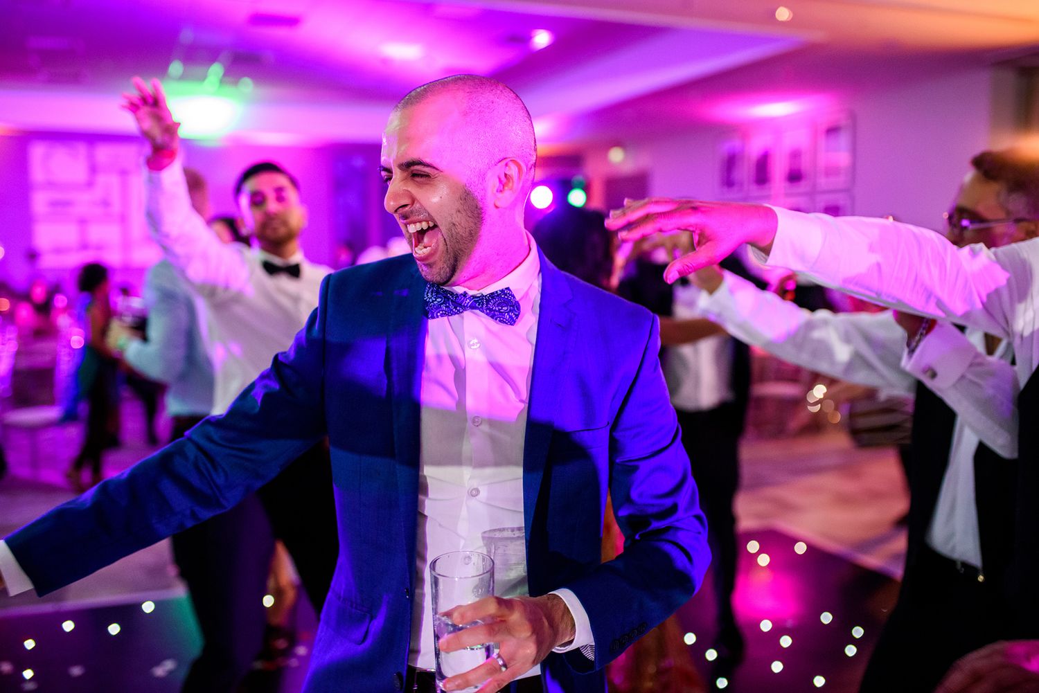 People dancing energetically at a wedding reception under dramatic purple lighting.