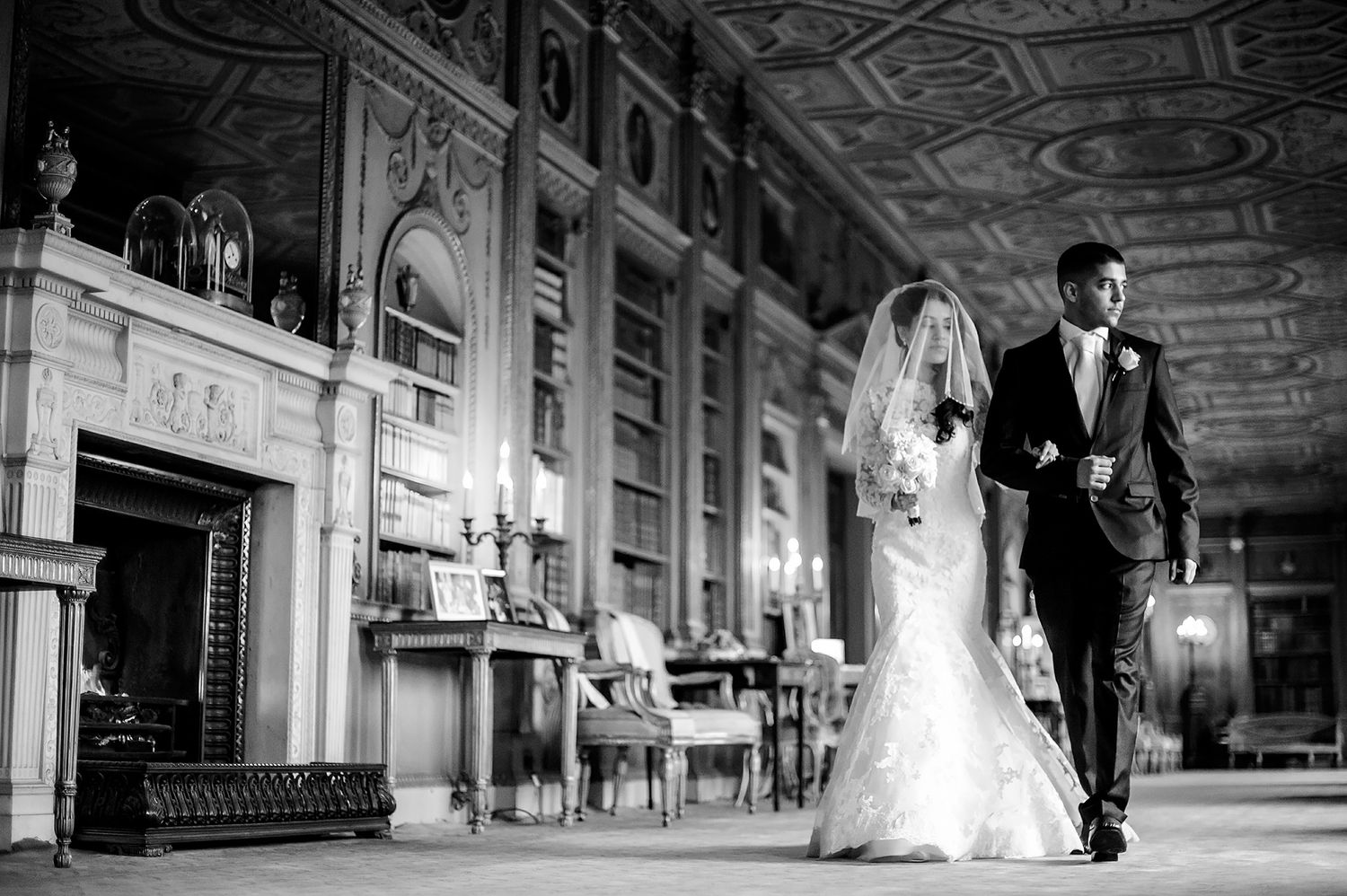 Black and white photo of a wedding couple walking through an ornate hallway with dramatic architectural details.
