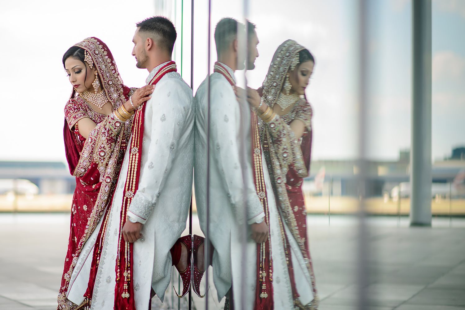 Wedding couple in traditional South Asian attire standing in modern glass building hallway.