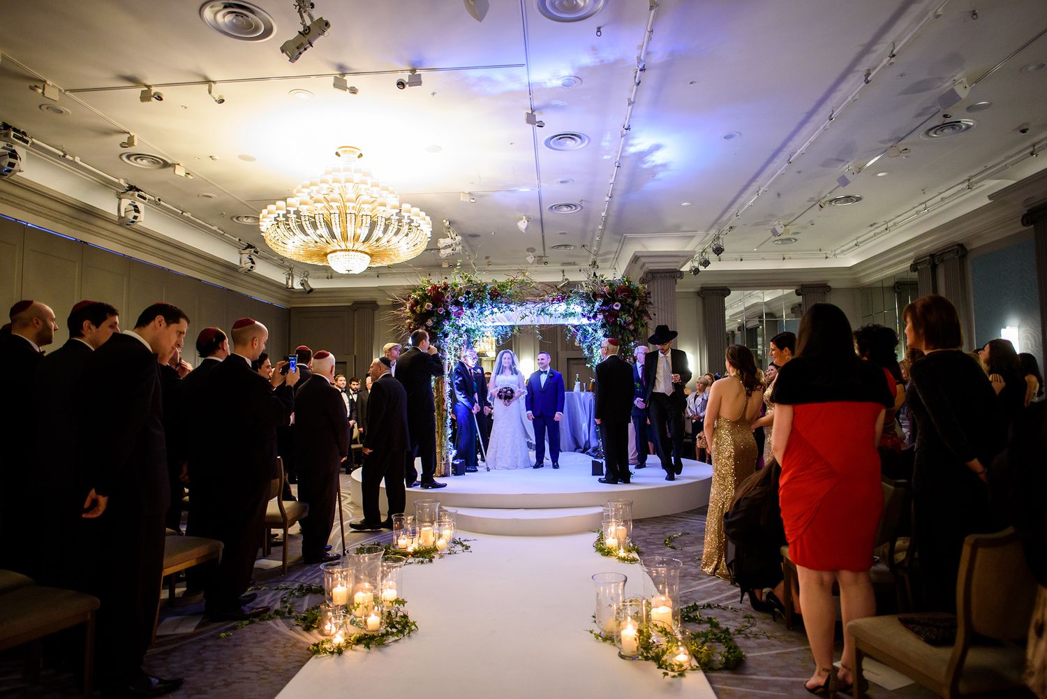 Wedding ceremony in progress with guests lining the aisle under elegant chandelier lighting.