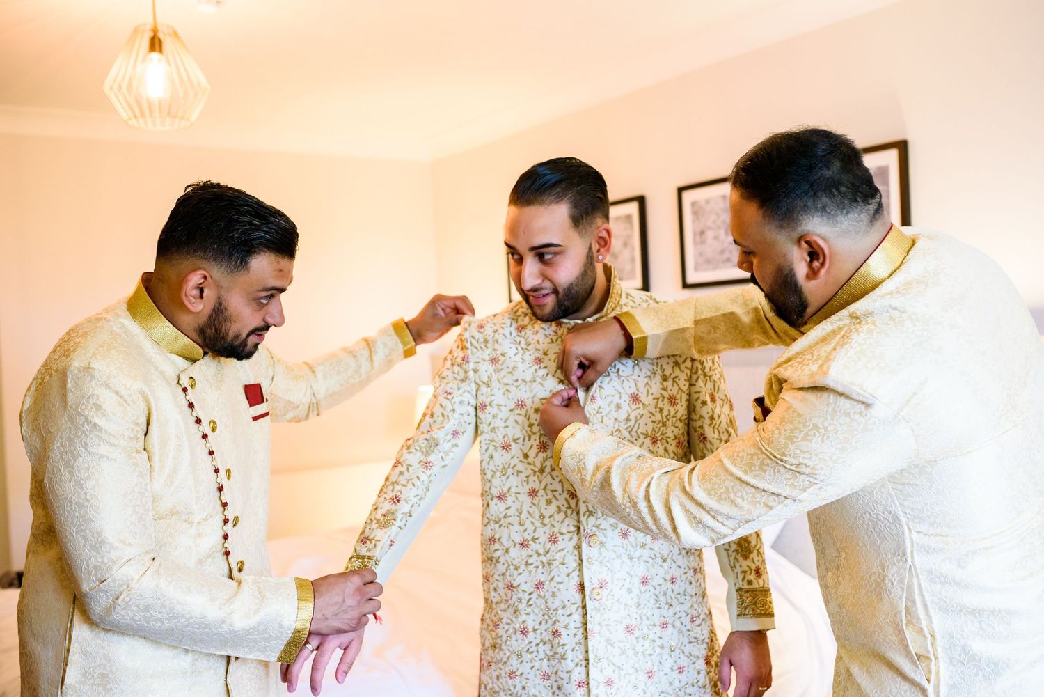 Men in traditional Indian wedding attire help each other get dressed in elegant cream-colored outfits.