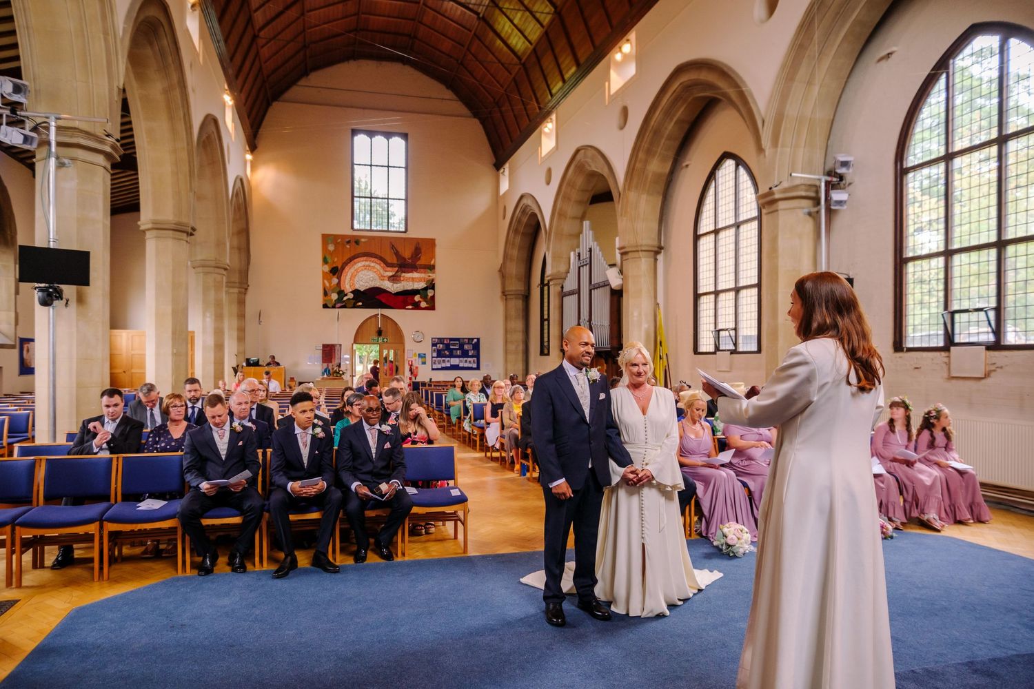 Panoramic view of a wedding ceremony in a traditional church with arched windows and guests seated in wooden pews.
