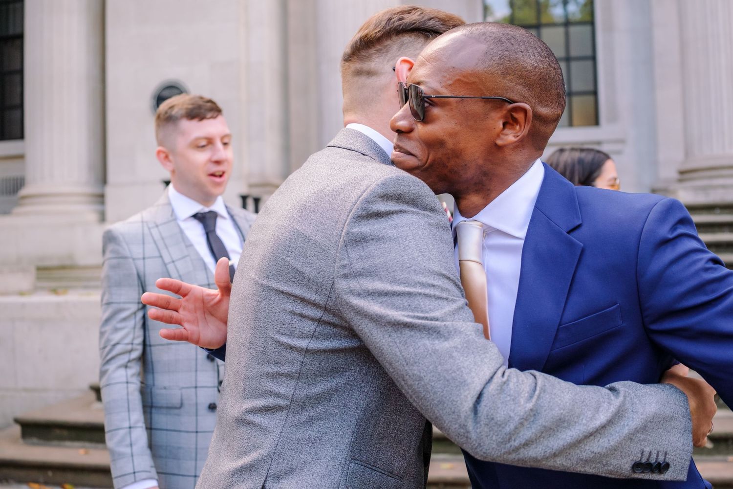 An emotional embrace between wedding guests outside a classical building.