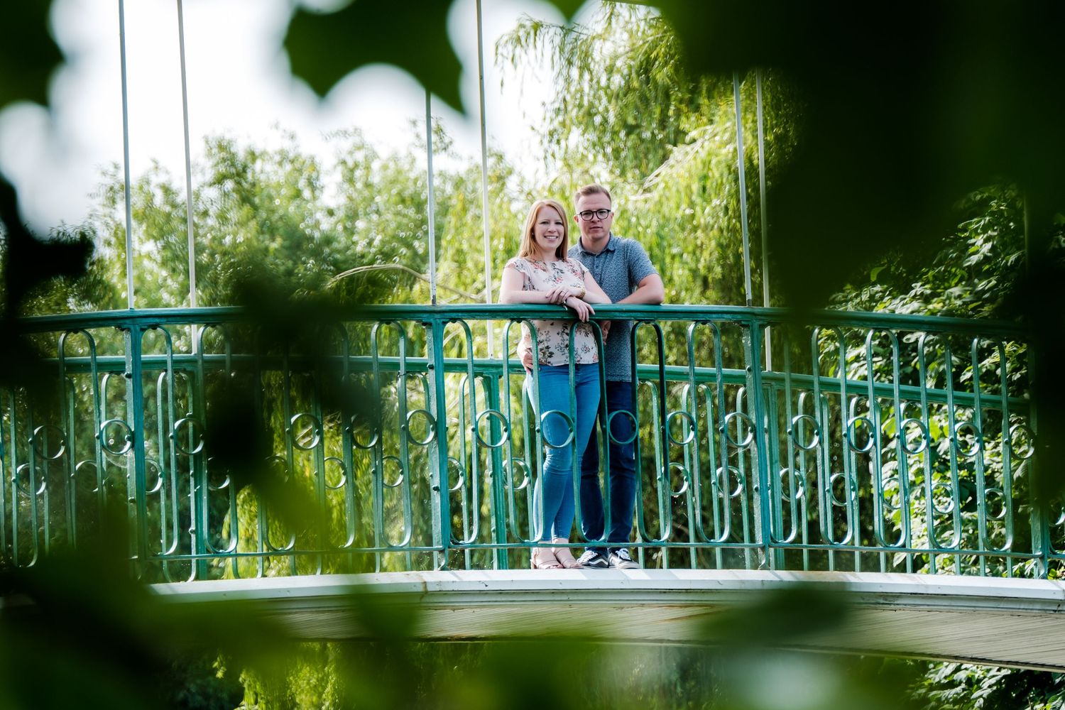 A couple stands on a turquoise bridge surrounded by lush greenery.