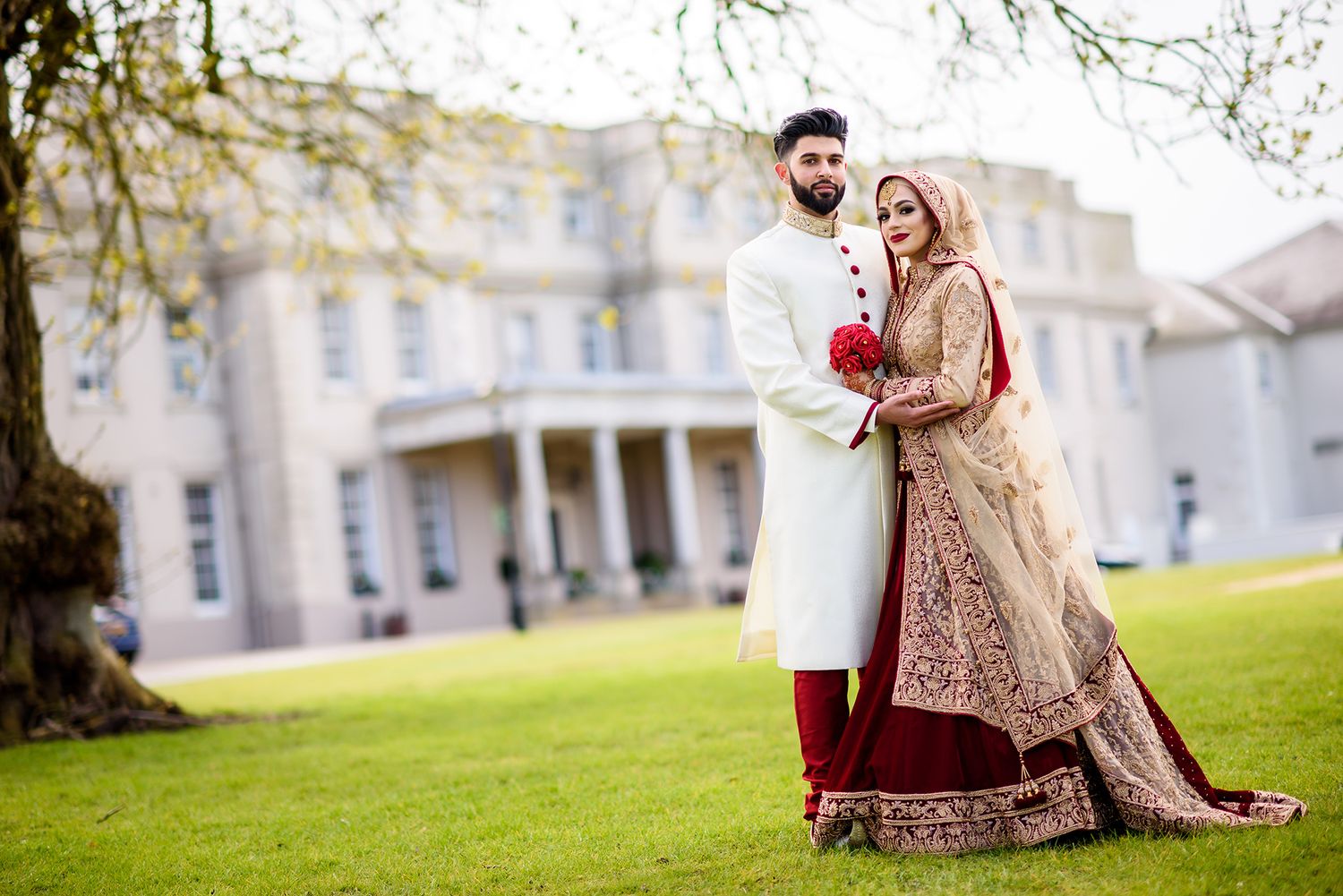 Wedding couple poses on manicured lawn with historic building in background.