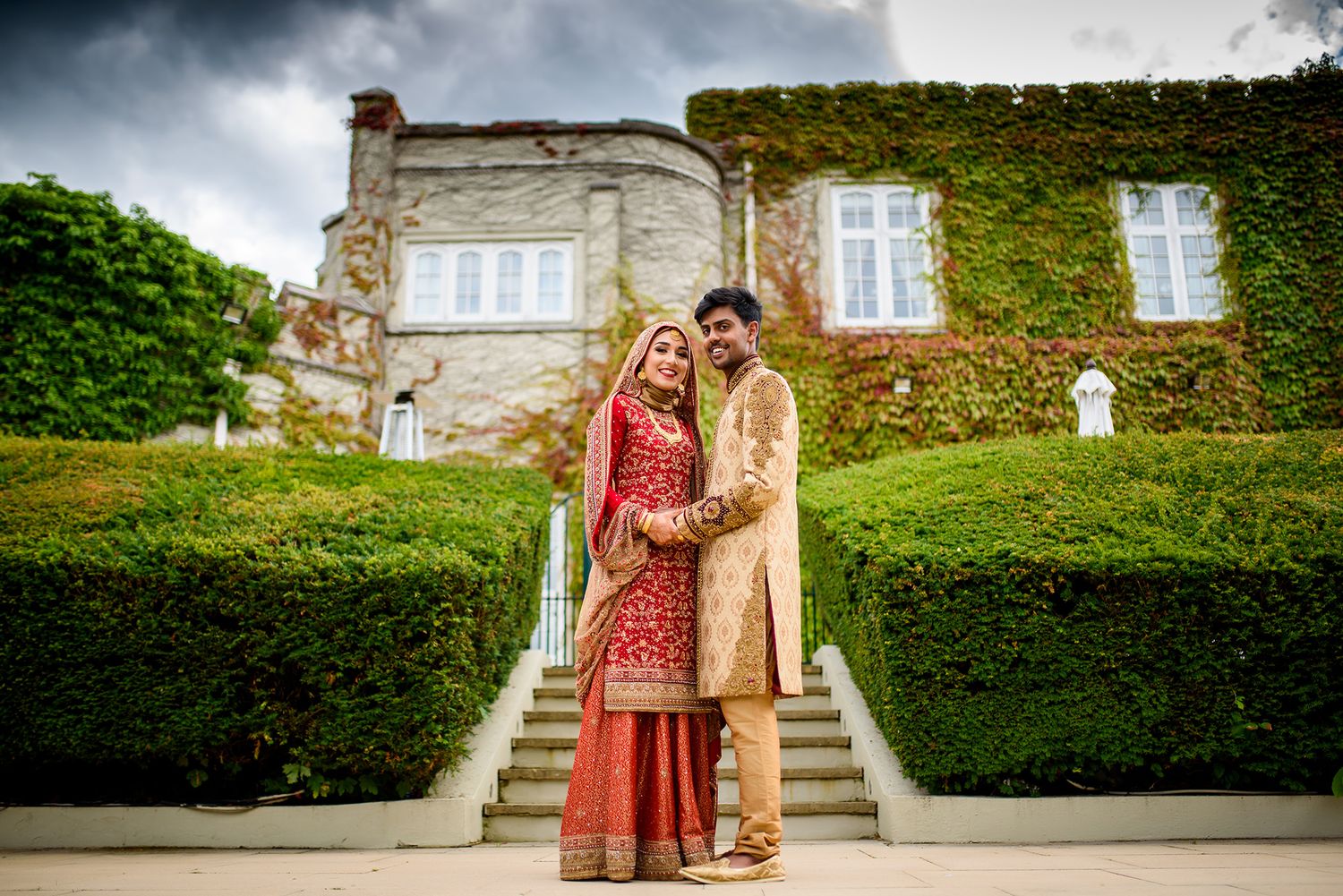 Wedding couple poses in traditional attire on grand stone steps in front of an ivy-covered building.