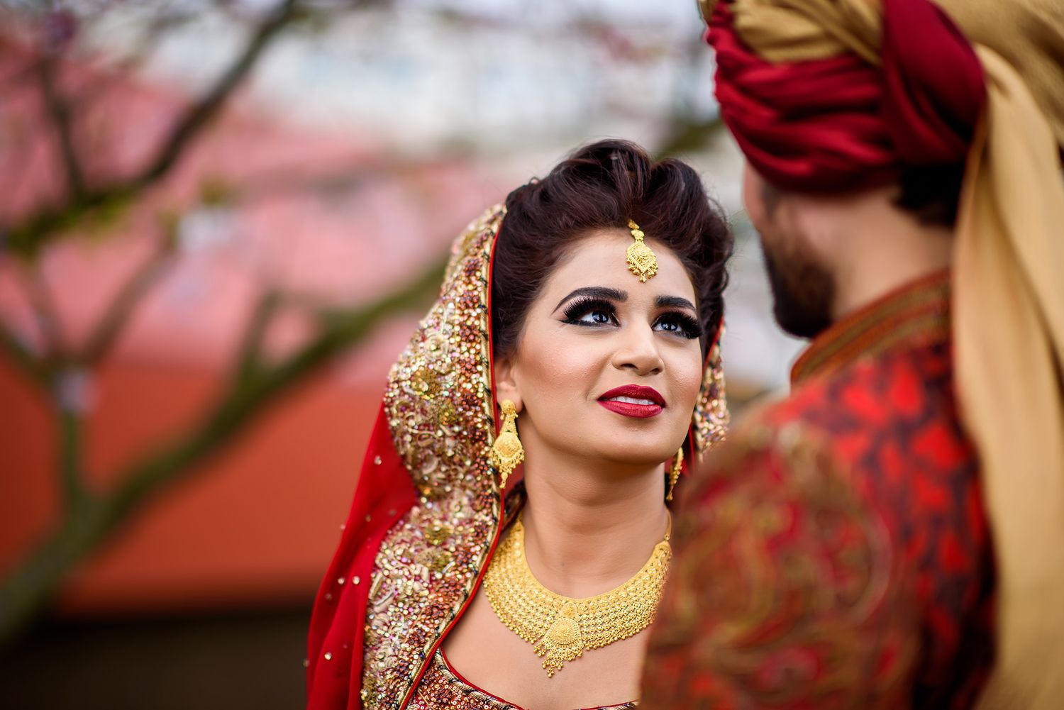 Couple in red and gold South Asian wedding attire pose under blooming cherry trees.