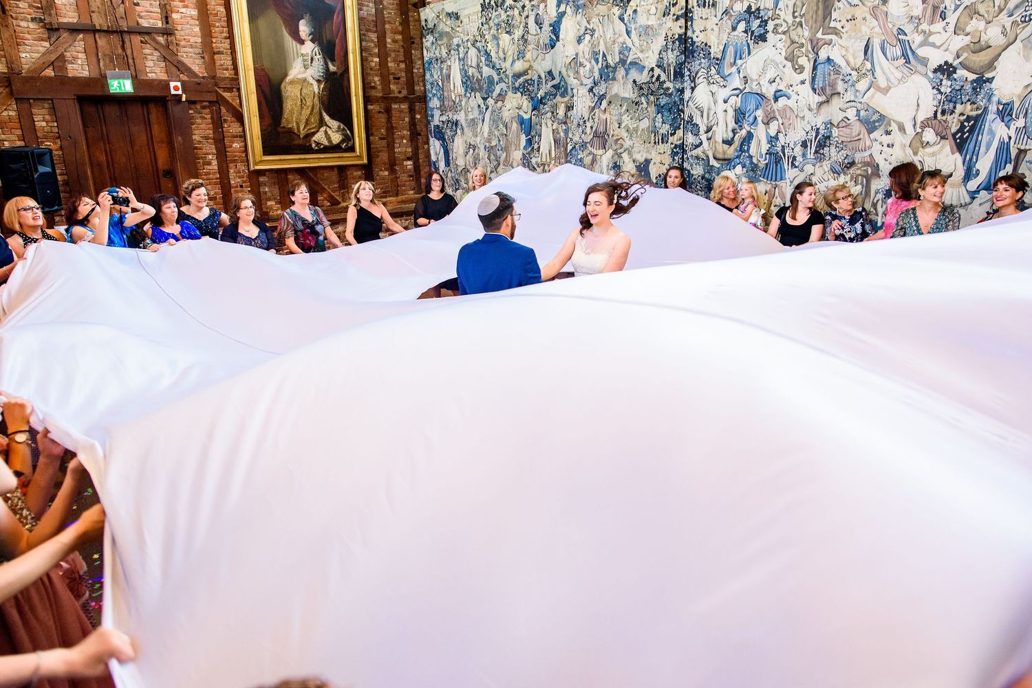 Wedding guests hold up a flowing white fabric canopy at an elegant indoor ceremony with decorative tapestries.