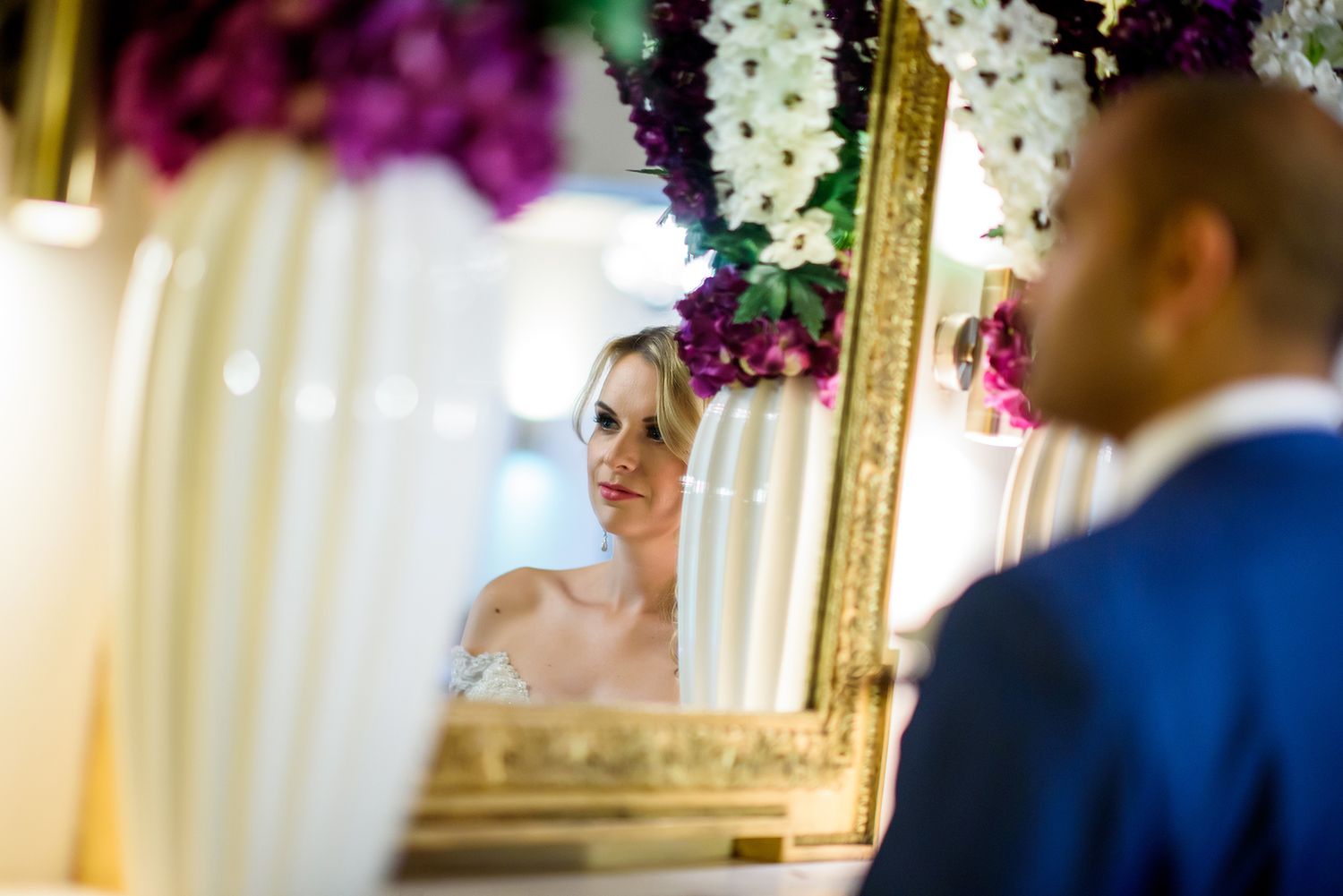 A reflection of the bride in an ornate gold mirror surrounded by purple and white flowers.