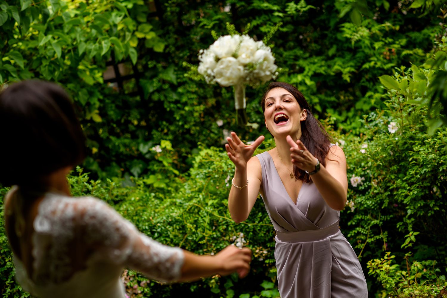 A person catches a flying wedding bouquet in a garden setting with joyful expression.
