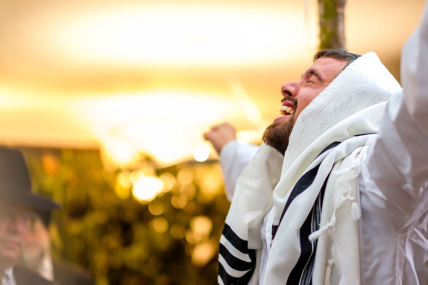 A couple wearing traditional Jewish prayer shawls exchanges rings during an outdoor sunset wedding ceremony.