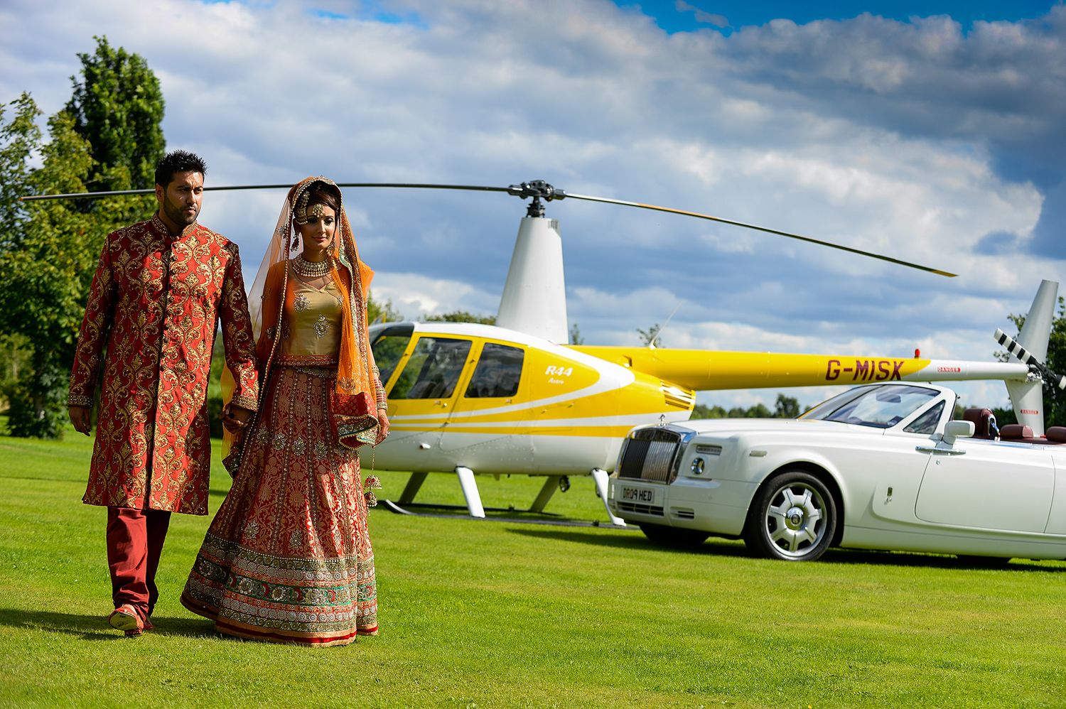 Couple in traditional Indian wedding attire standing near yellow helicopter and luxury car.
