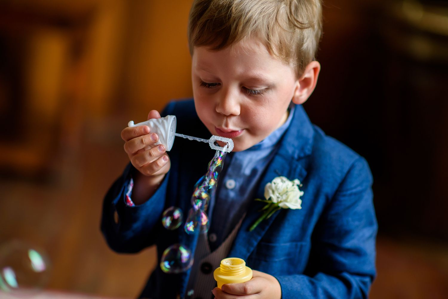 Young boy in blue suit blows bubbles during wedding celebration.