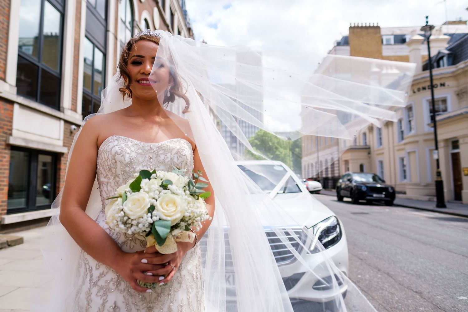 A person in a white wedding gown holds a bouquet of white flowers on a city street.