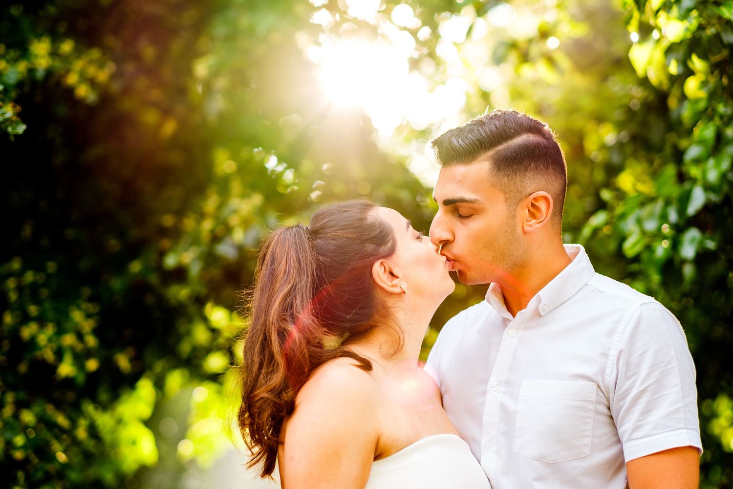 Couple sharing romantic kisses in a sun-drenched garden setting with green foliage in the background.
