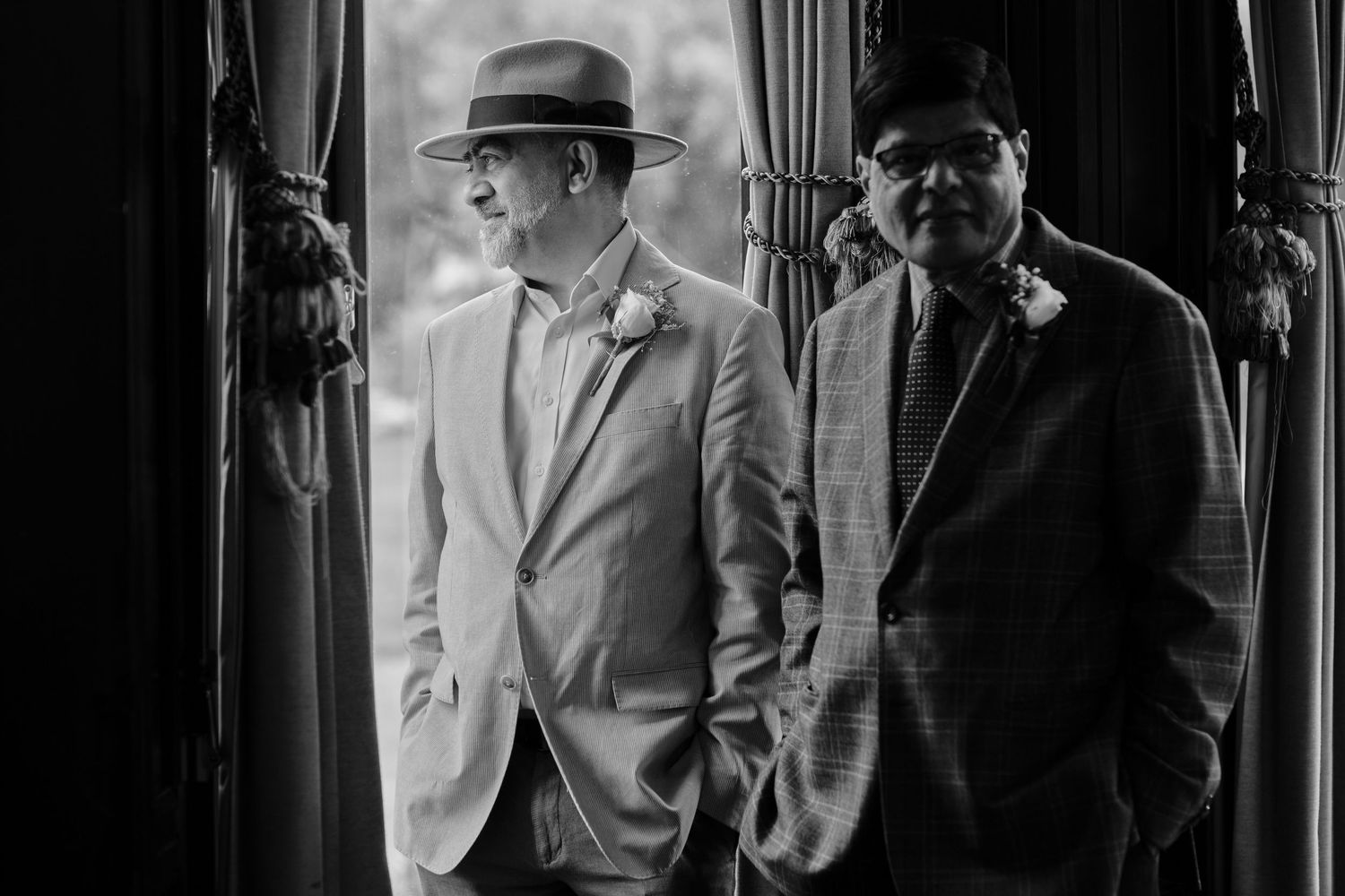 Two men in formal suits and hats stand together in dramatic black and white lighting.