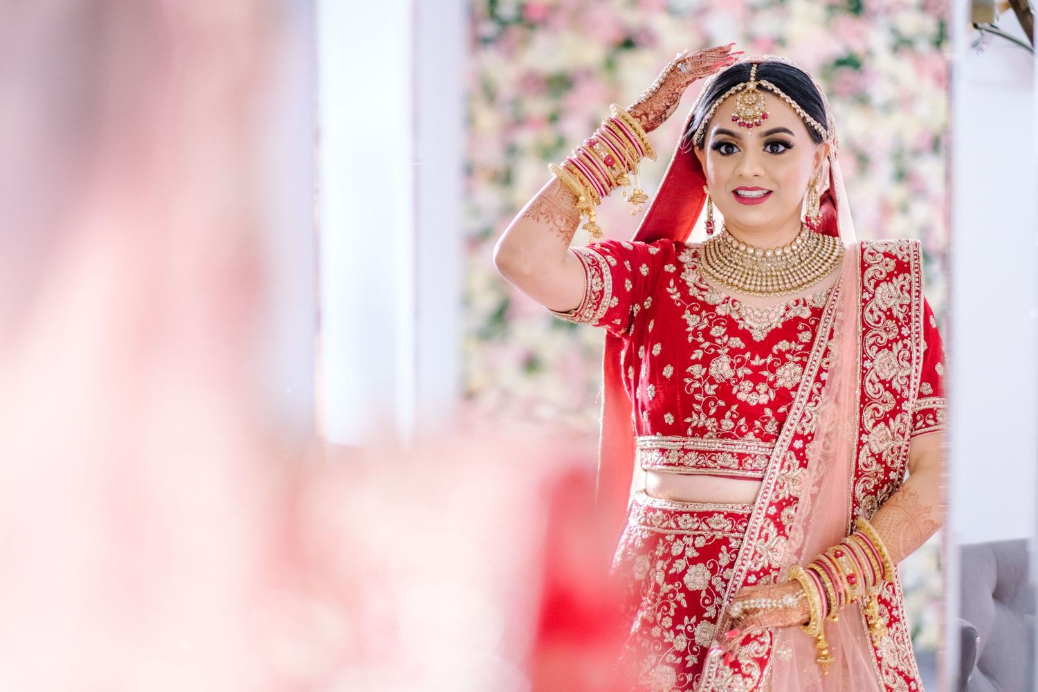 A traditional Indian bride wearing a red and gold lehenga with ornate jewelry and bridal accessories in a bright setting.