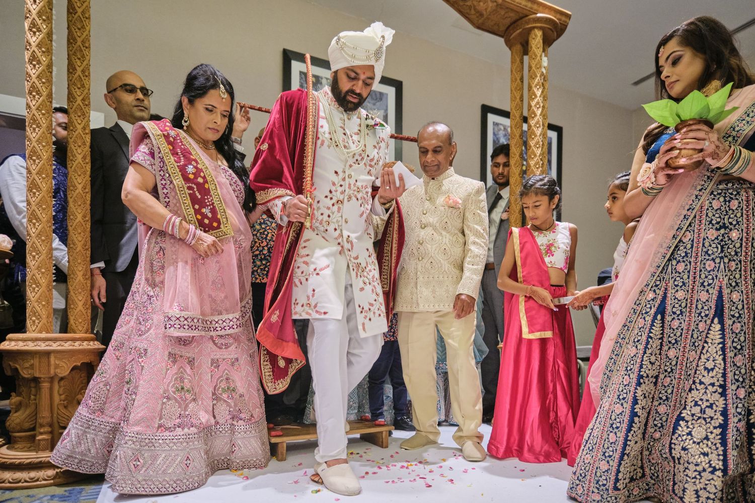 A traditional Indian wedding ceremony with guests in colorful lehengas and formal attire gathered around decorative mandap pillars.