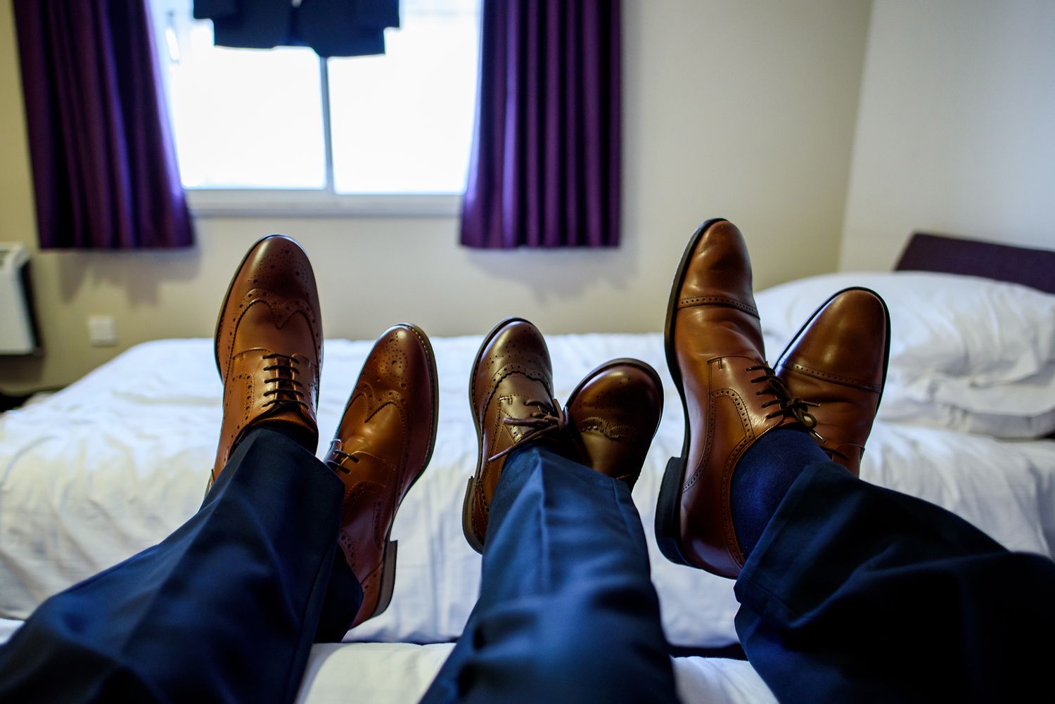 Brown leather dress shoes resting on bed with blue pants visible.
