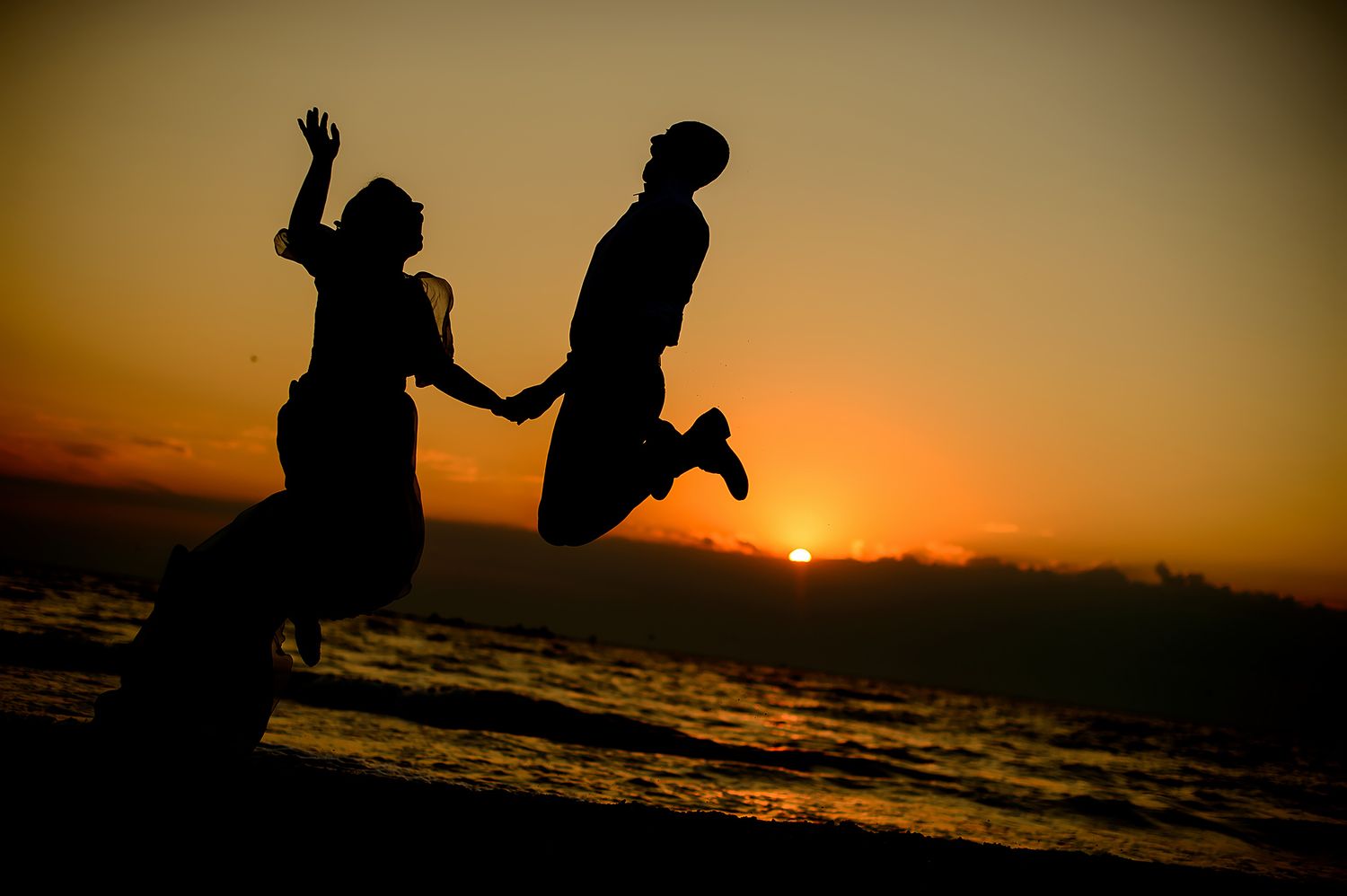 Silhouetted couple jumping together against a dramatic sunset over the ocean.
