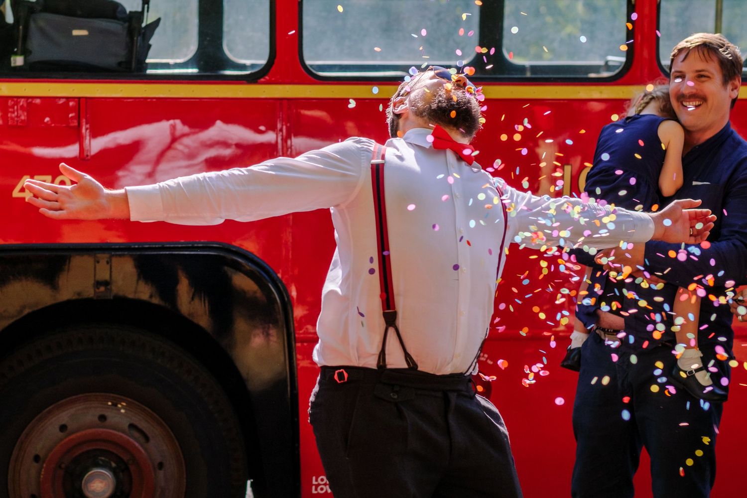 A festive celebration scene with white confetti falling in front of a red bus or vehicle.