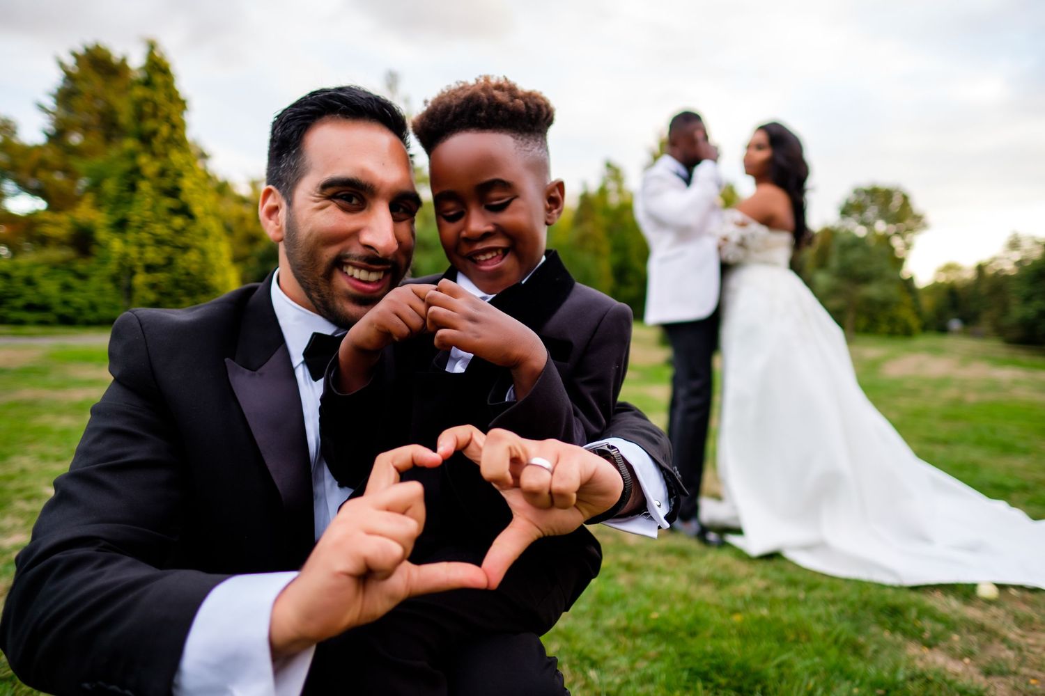 Groomsmen making heart shapes with hands on grassy lawn during wedding.