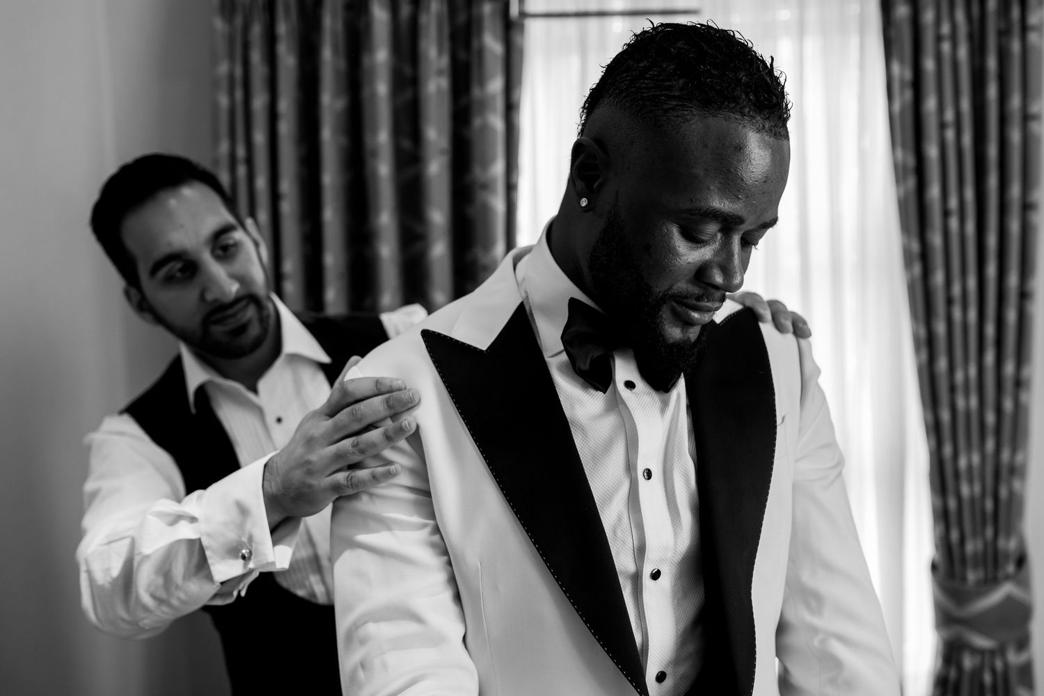 Black and white image of groomsmen helping each other prepare in formal wear before a wedding.