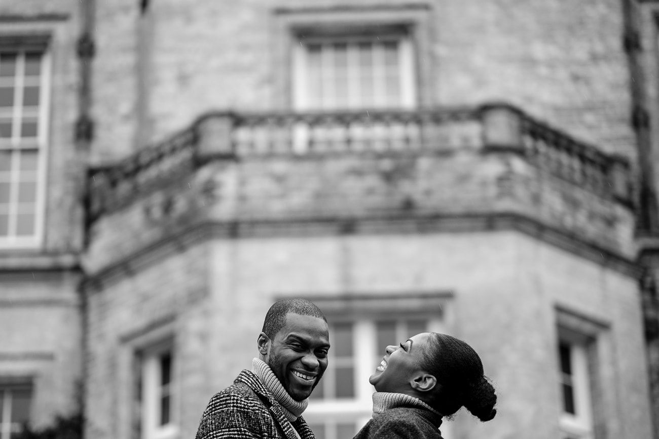 A romantic couple laughing together in front of a historic stone mansion.