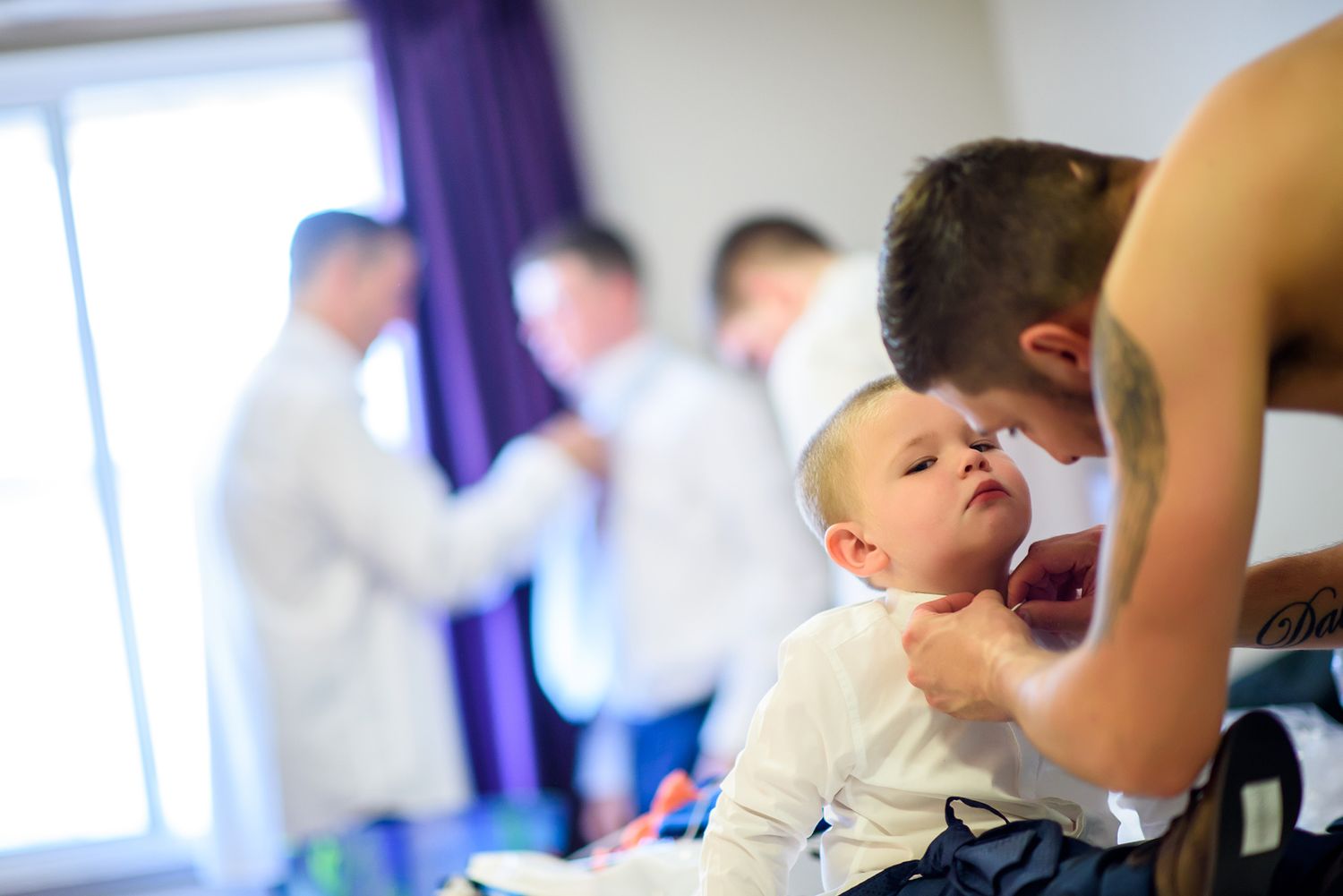 A child getting dressed in formal attire with others in the background of a room with purple curtains.