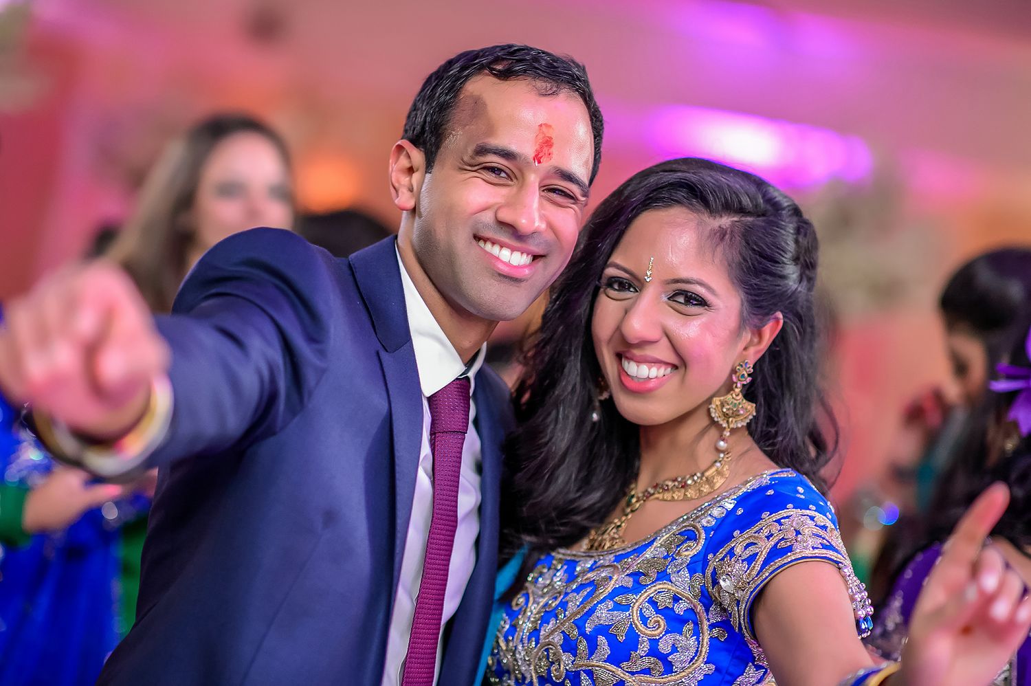 Wedding celebration in a vibrant pink-lit venue with guests in formal Indian attire.