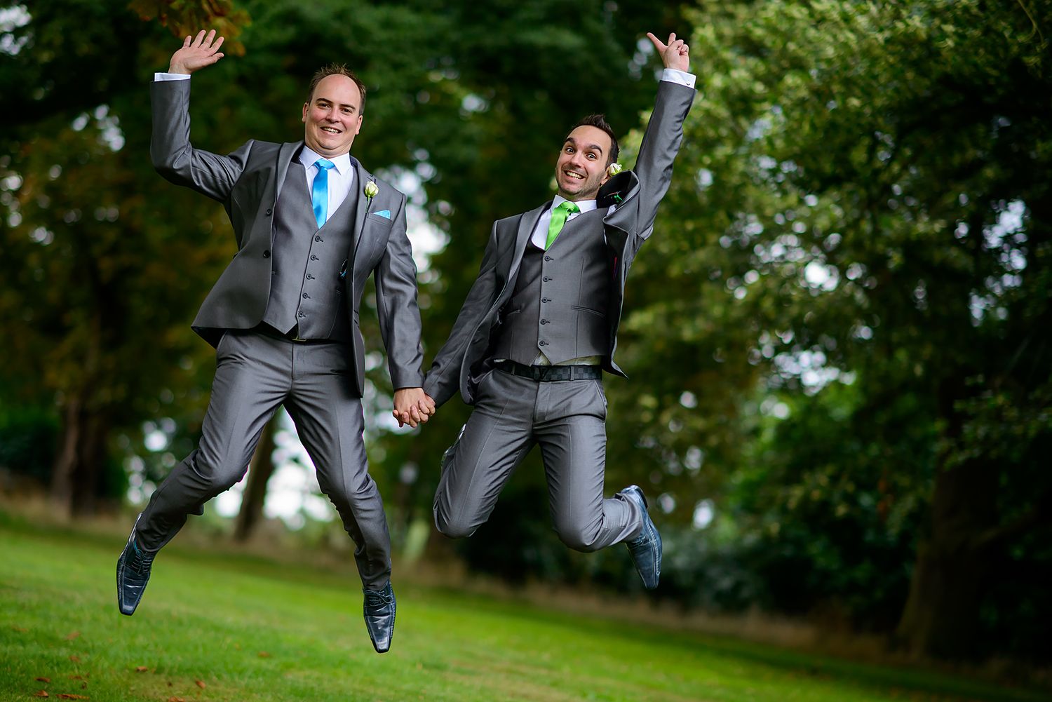 Joyful couple in matching gray suits jump together on grass outdoors.