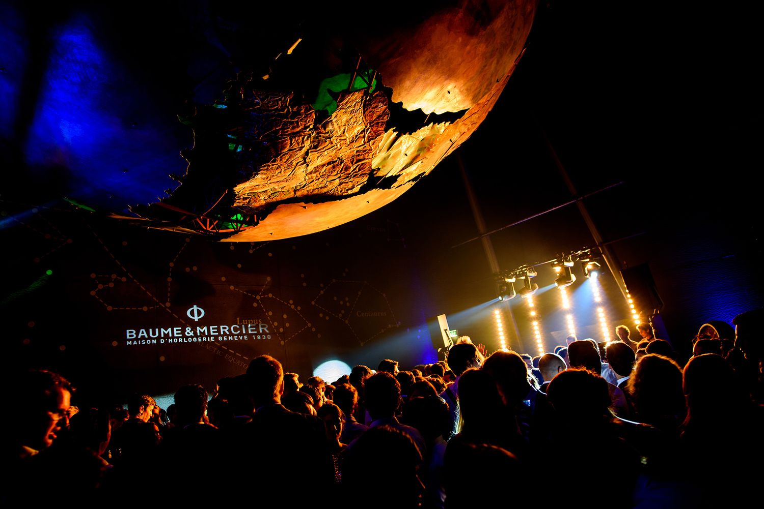 A dark concert venue with a silhouetted crowd beneath colorful stage lighting and illuminated screens.