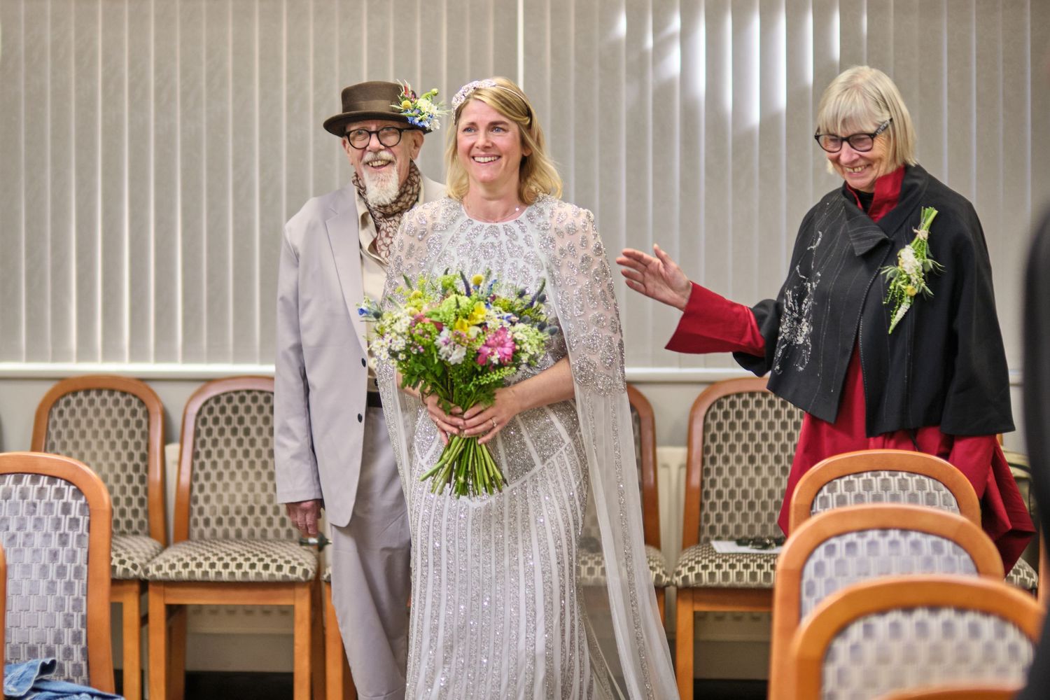 Wedding ceremony in a bright room with white vertical blinds and wooden chairs.