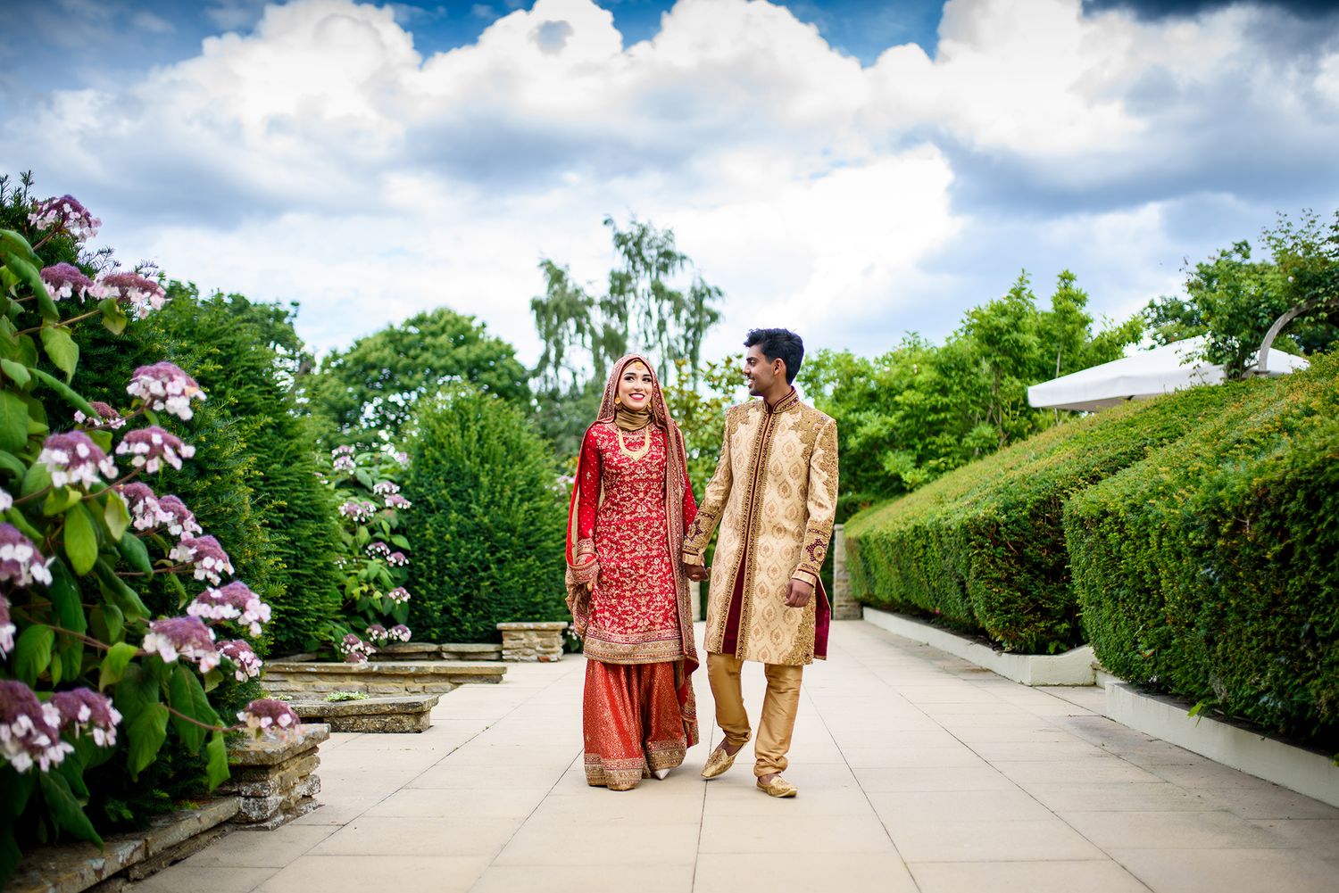 Wedding party walking through landscaped garden path with blooming flowers and cloudy sky.