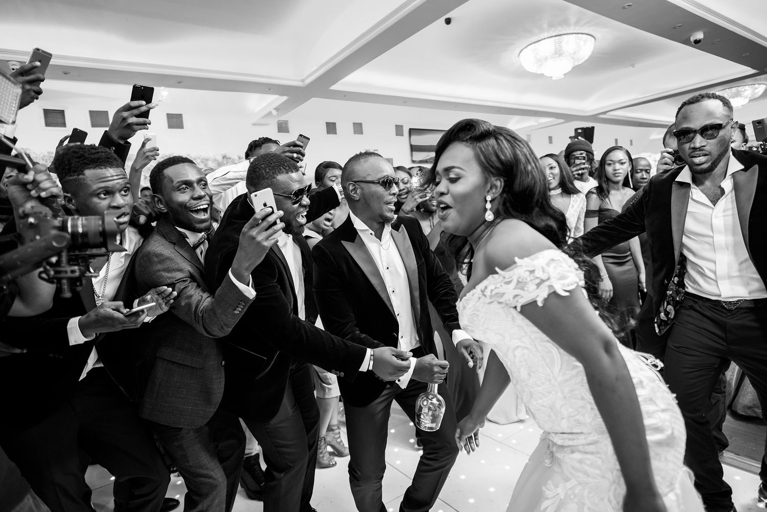 Wedding celebration with bride and guests dancing joyfully in a reception hall captured in black and white.