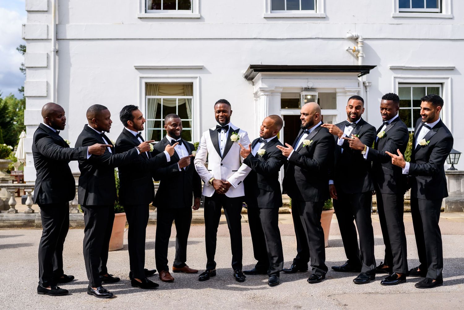 Groomsmen in dark suits pose outdoors in front of a white building during a wedding celebration.
