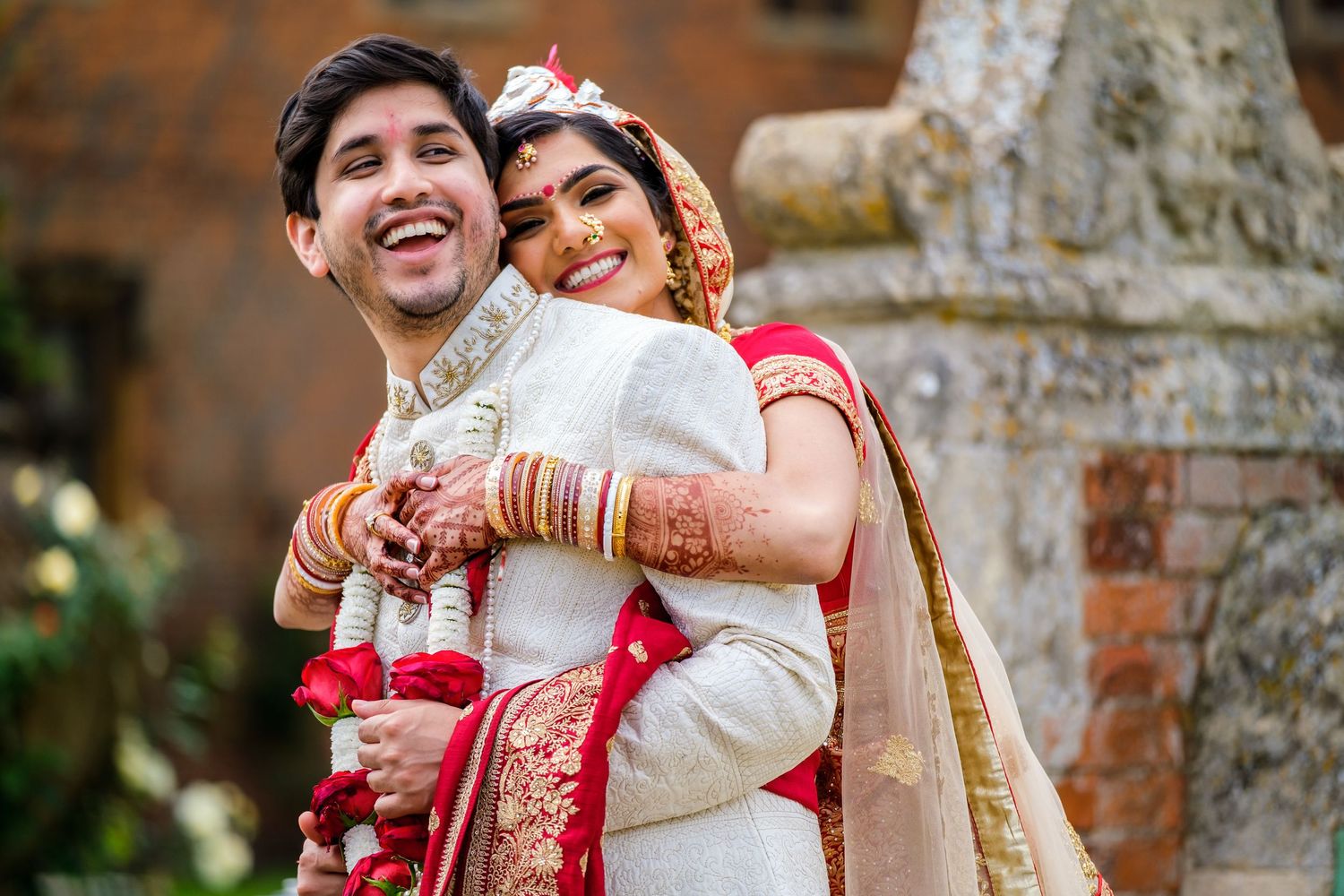 Joyful wedding portrait featuring traditional Indian bridal wear against historic brick backdrop.