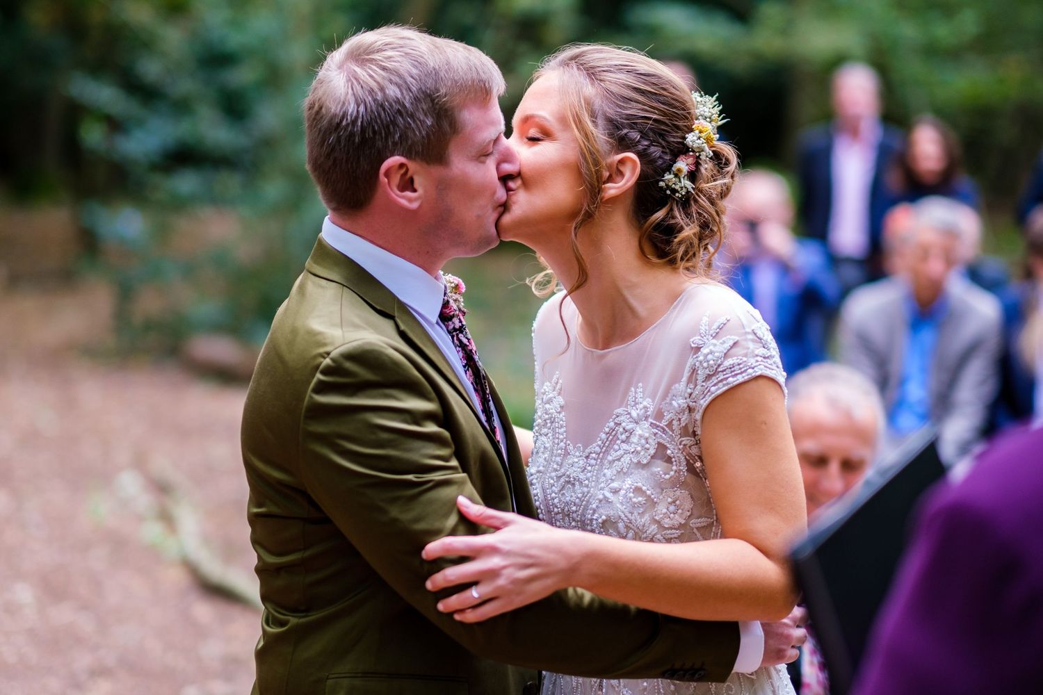 Romantic wedding ceremony kiss in an outdoor woodland setting with guests watching.