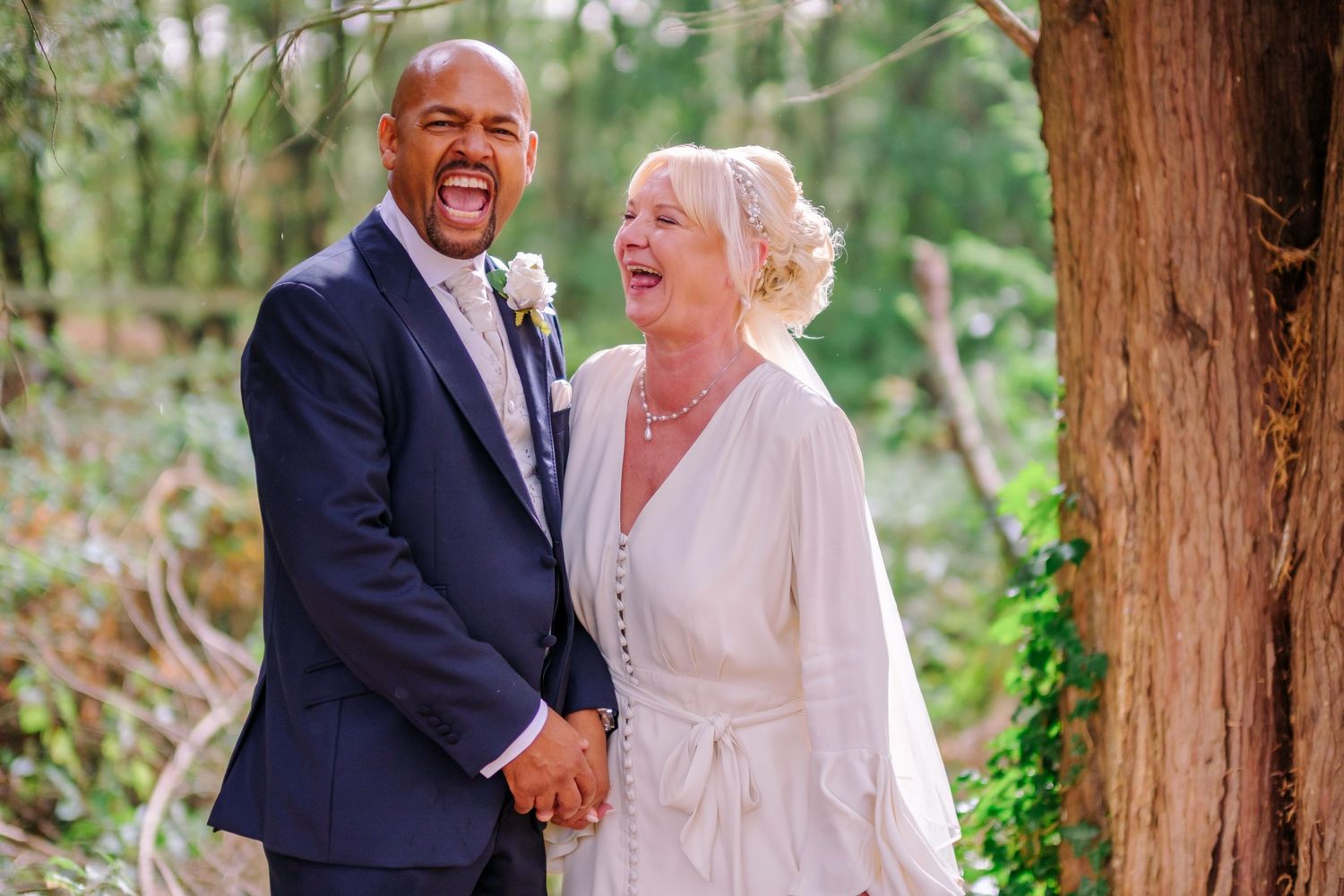 A joyful couple shares a candid moment laughing together outdoors among tall trees during their wedding celebration.