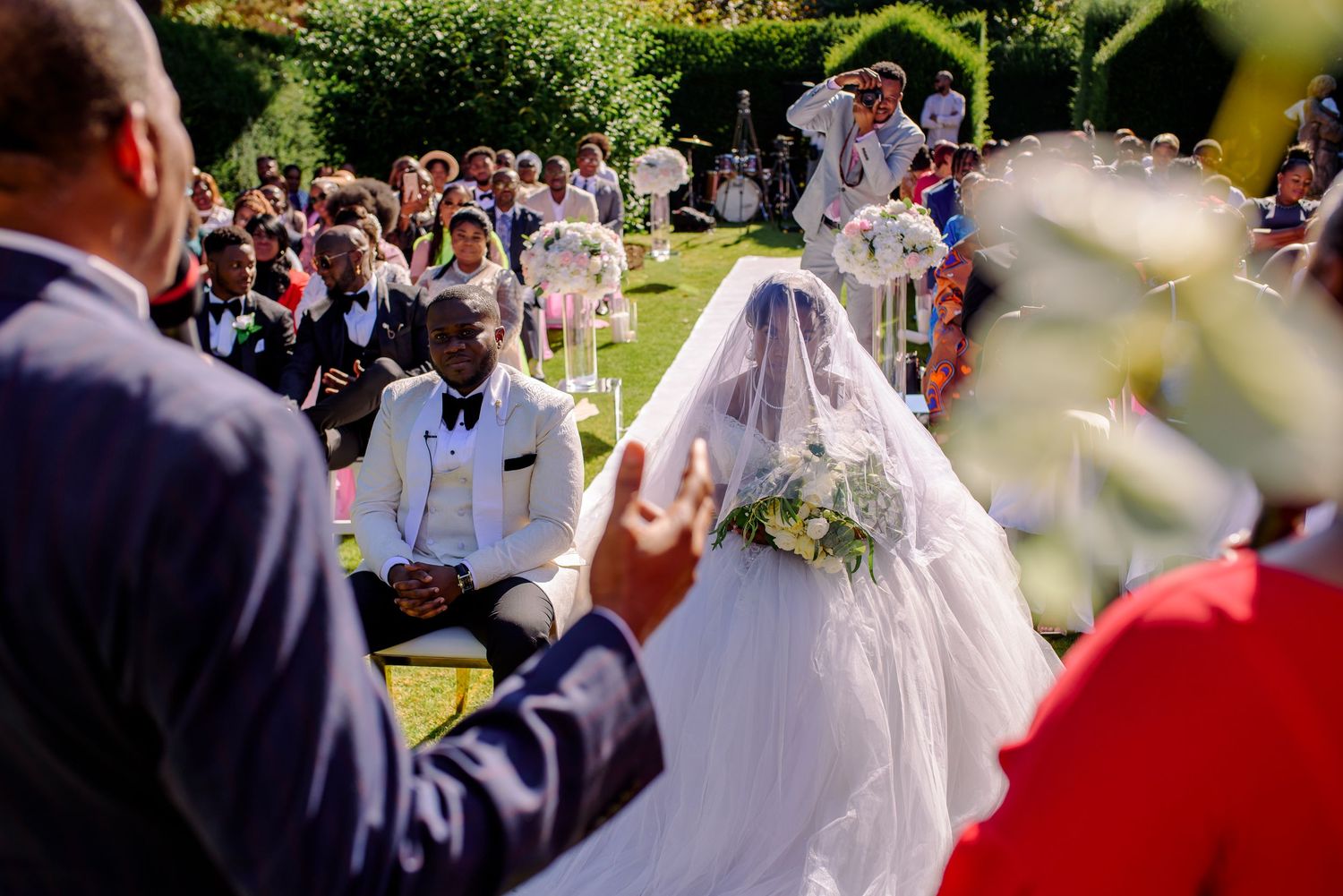 Wedding ceremony takes place in a lush garden setting with guests seated on white chairs.